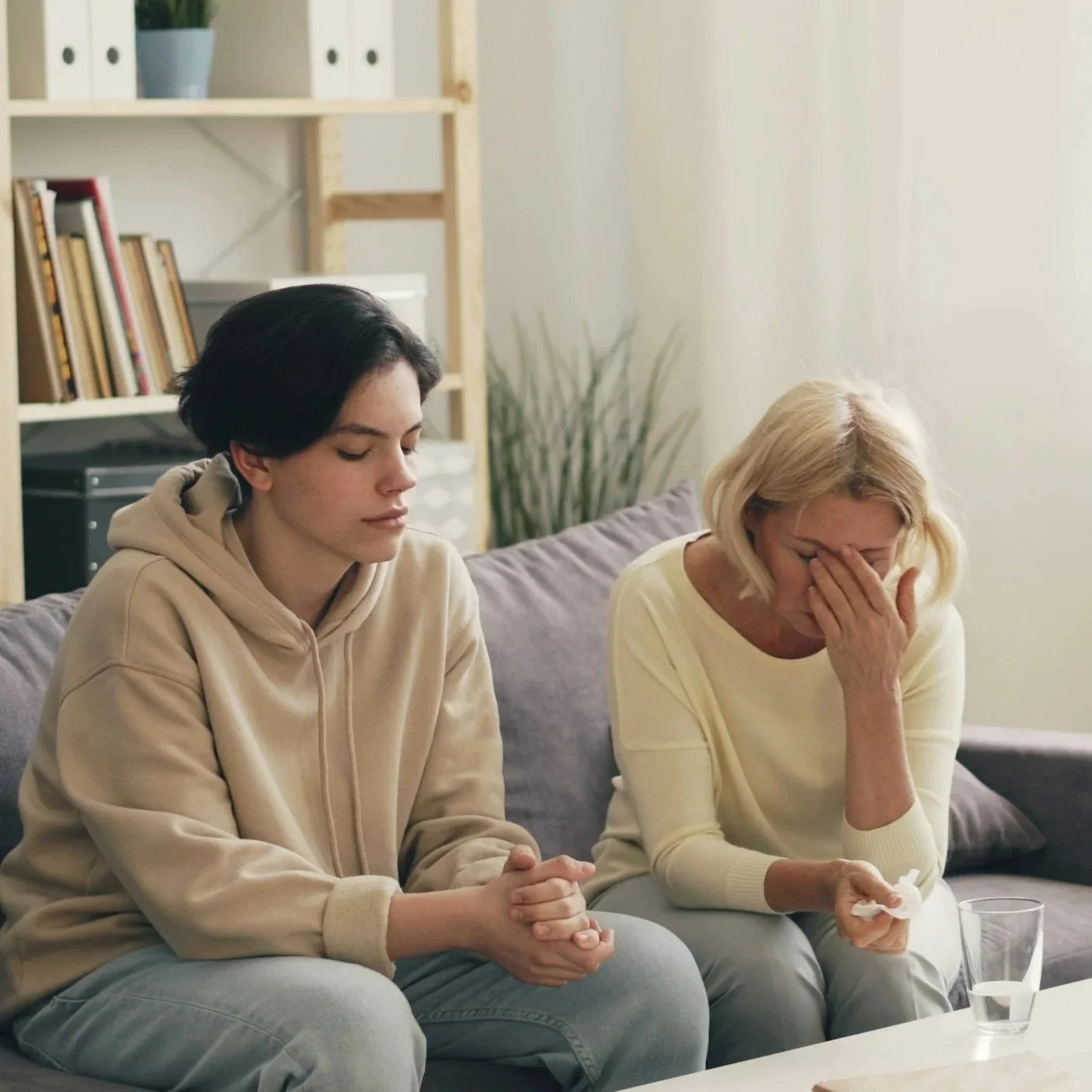 A young woman and an older woman sit on a gray couch in a living room, both with eyes closed and emotional expressions; the young woman is wearing a beige hoodie, and the older woman is wearing a light yellow sweater, covering her face with her hand.
