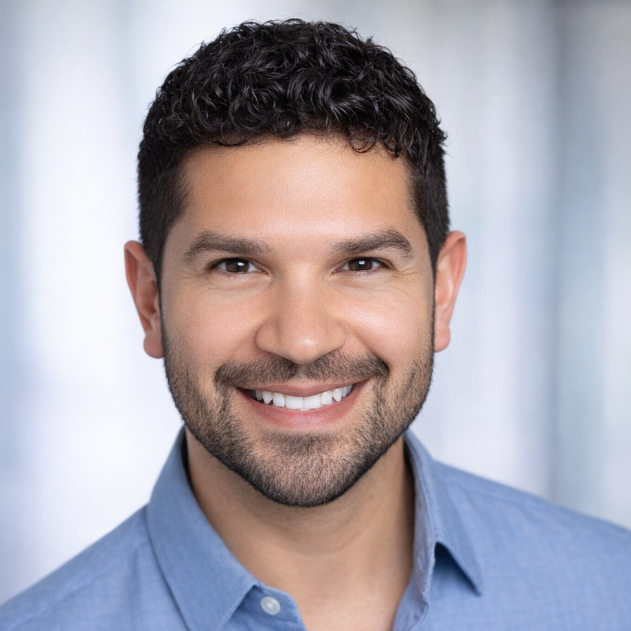 Headshot of a smiling man with dark, curly hair, wearing a light blue collared shirt.