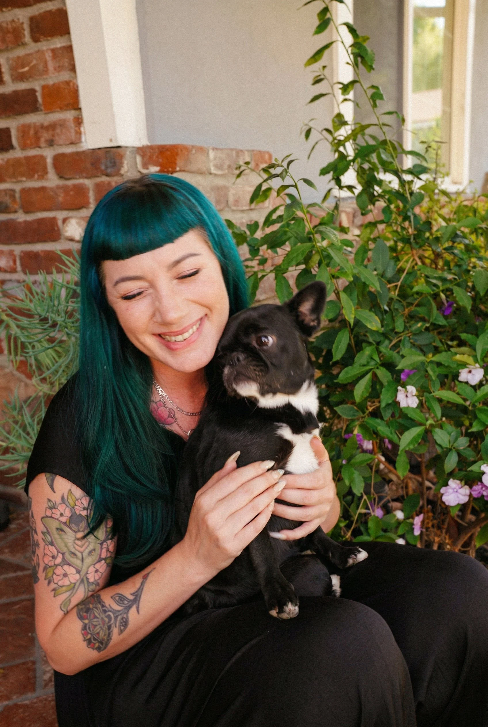 A woman with teal hair and tattoos on her arm is smiling while holding a black and white puppy. They are sitting outside near a brick wall and lush green plants with purple flowers, near a window.