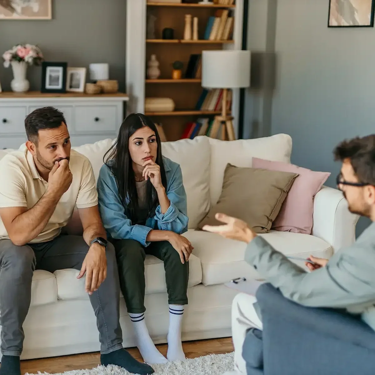 A therapy session with two clients sitting on a cream sofa and a counselor sitting on a blue chair in a living room with bookshelves and decorated wall art.