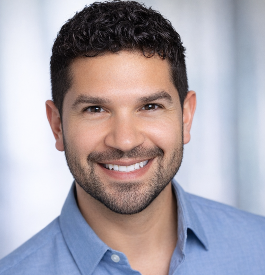 A smiling man with dark curly hair and a beard wearing a light blue button-up shirt, standing indoors with a blurred light background.
