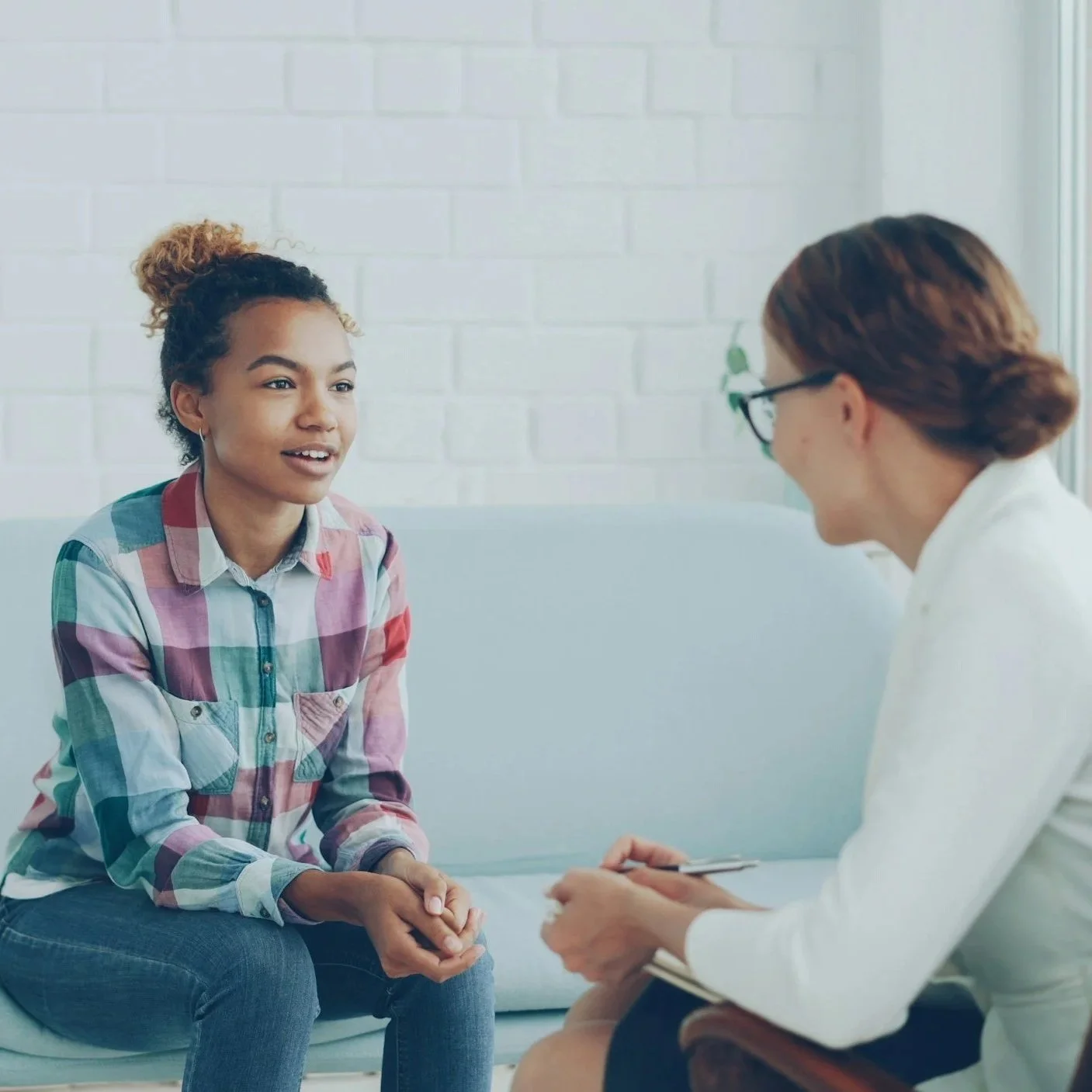 A young woman with curly hair and a plaid shirt sitting on a white couch, talking to a female therapist or counselor with glasses, a white shirt, and a notebook.