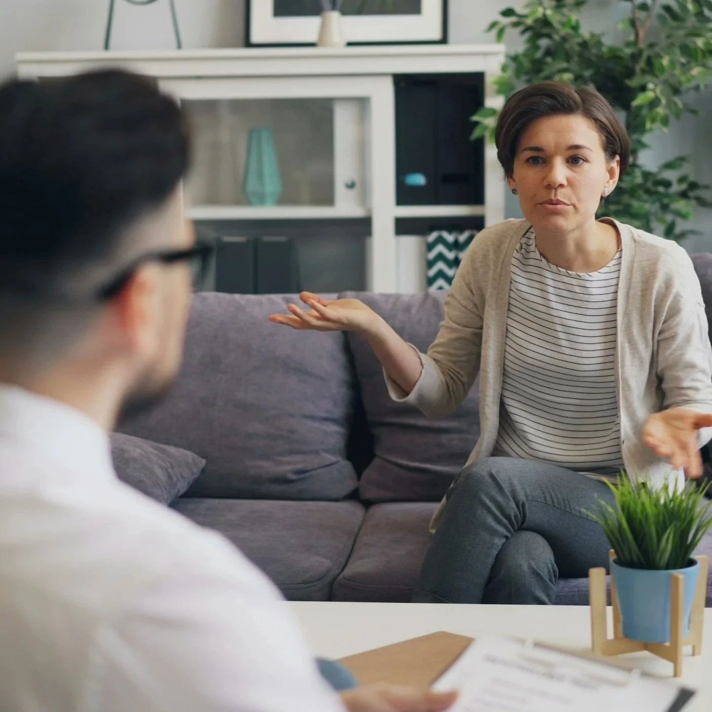 A woman with short dark hair gesturing with her hands while talking to a person with glasses in a cozy living room.