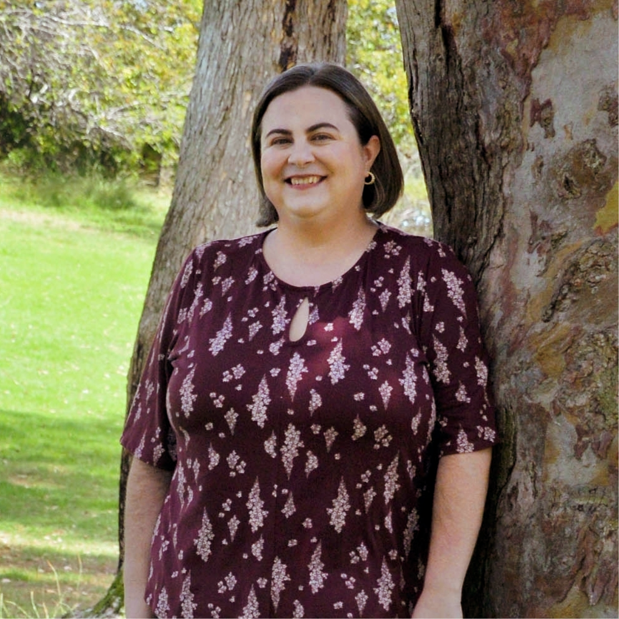 A woman with shoulder-length dark hair wearing a burgundy dress with white floral patterns, standing outdoors next to a large tree with textured bark, smiling at the camera.