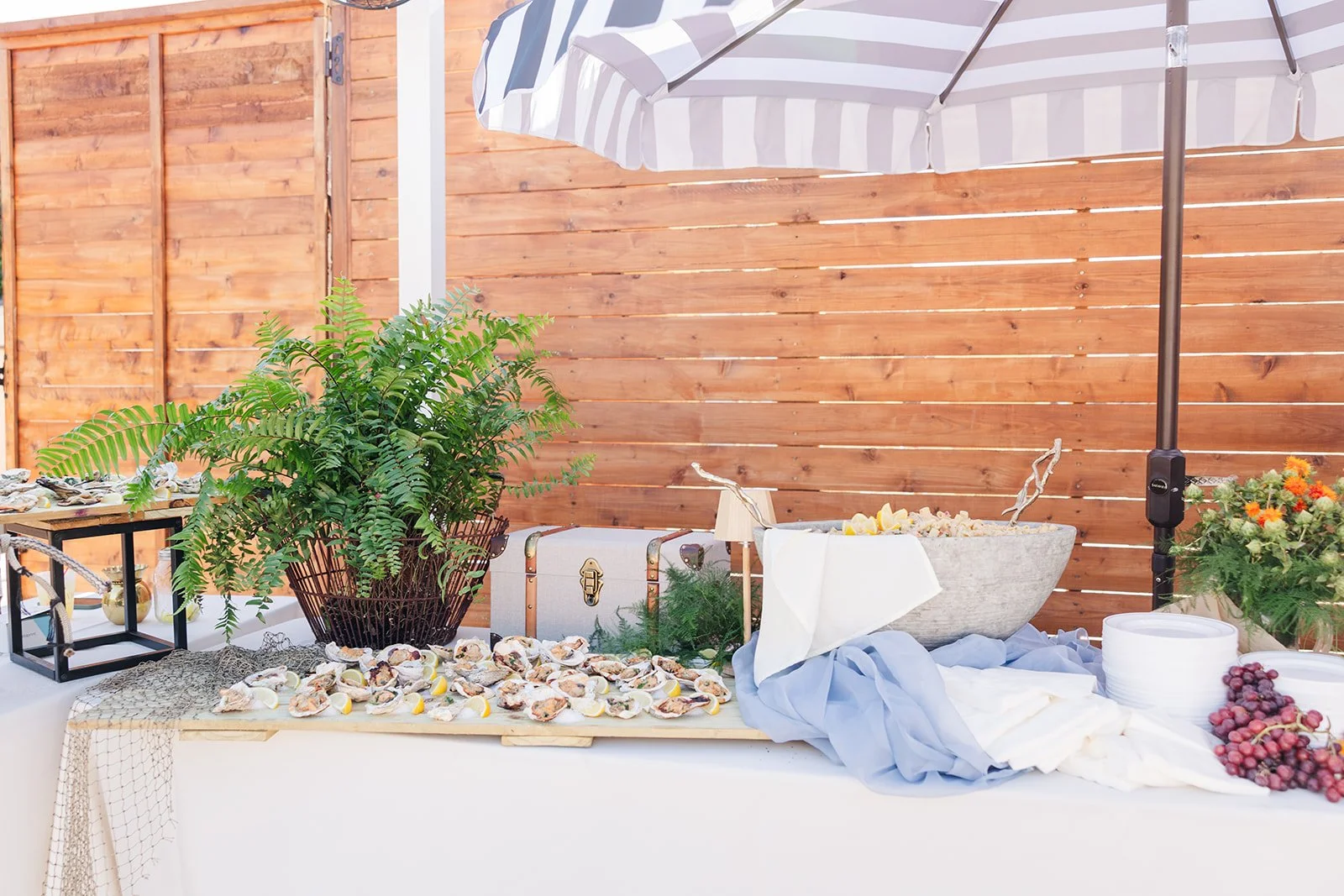 A table with seafood and lemon slices, decorated with plants and a large umbrella, set against a wooden fence.