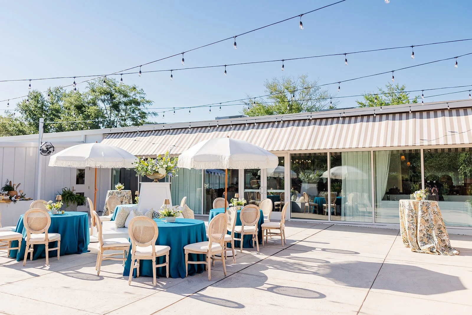 Outdoor event setup with round tables covered in blue tablecloths, surrounded by beige chairs, white umbrellas, string lights overhead, and a glass-walled building in the background.