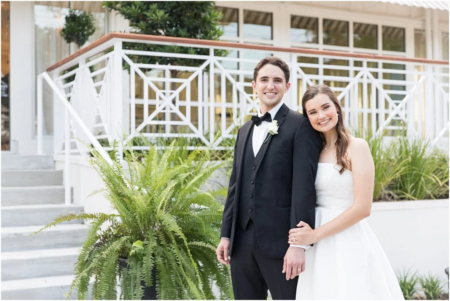 A newlywed couple smiling, standing outdoors near stairs and green plants, with a white railing and a building in the background.