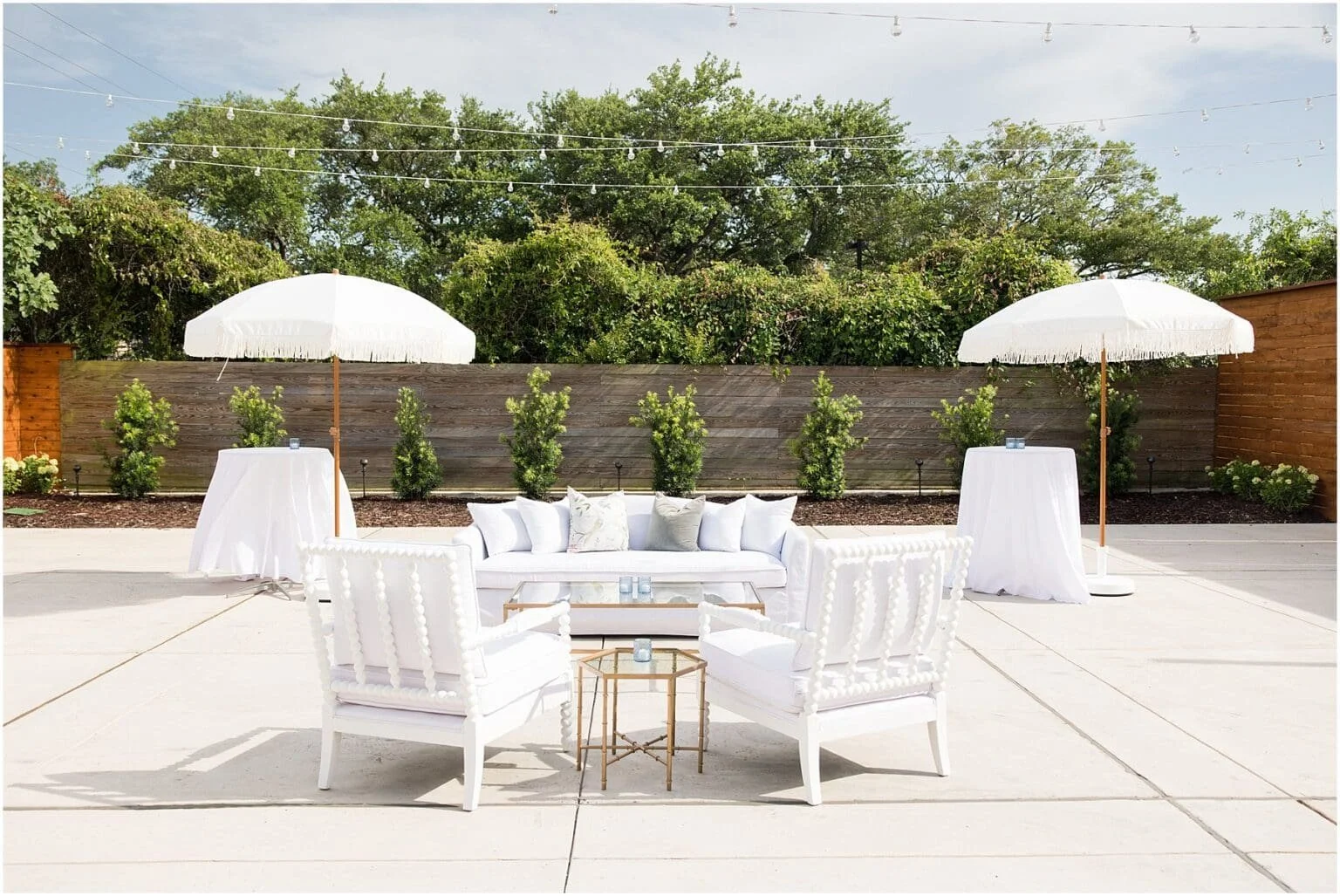 Outdoor seating area with white furniture, two large white umbrellas, and string lights, set on a concrete patio with greenery and trees in the background.