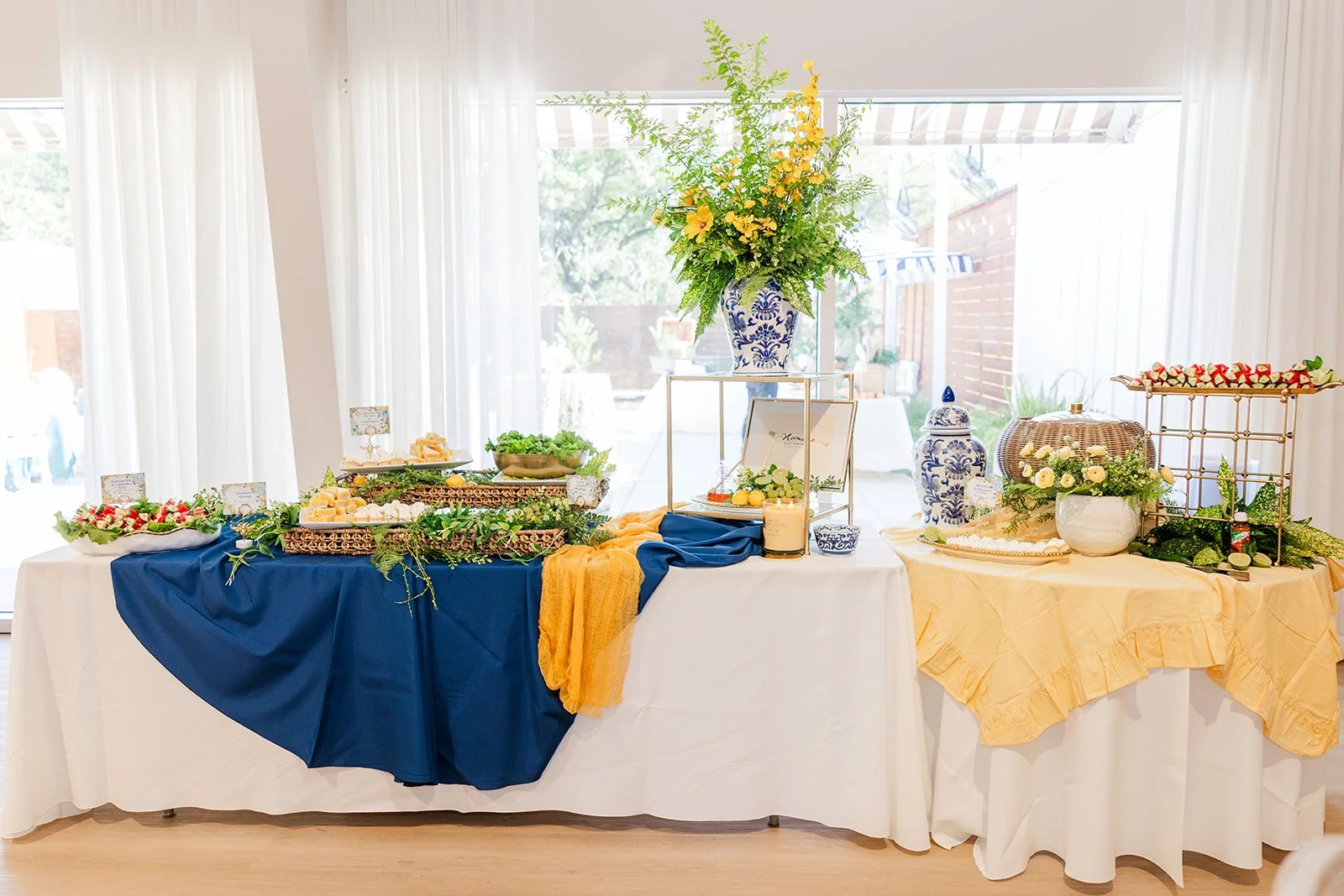 Buffet table with various dishes and decorations, including a tall floral arrangement in a blue and white vase, covered with blue and yellow tablecloths, set against white curtains.