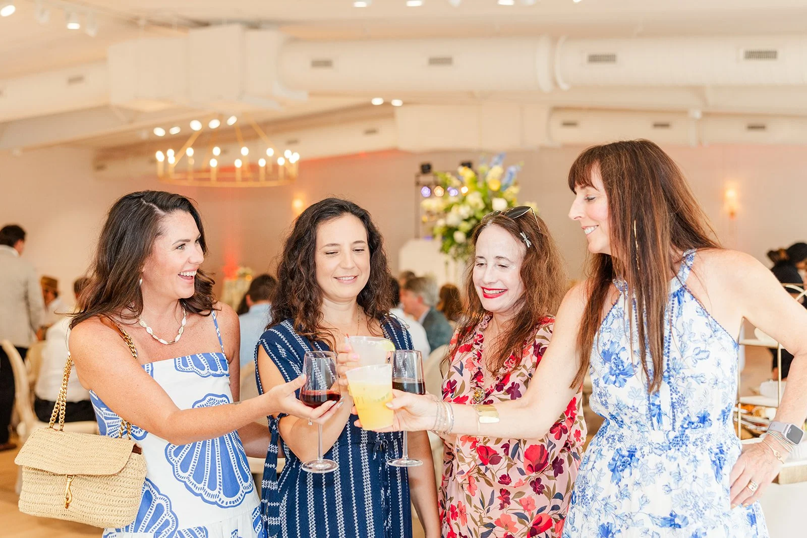 Four women standing together at a social event, holding glasses of red wine and a yellow cocktail, smiling and clinking glasses in celebration.