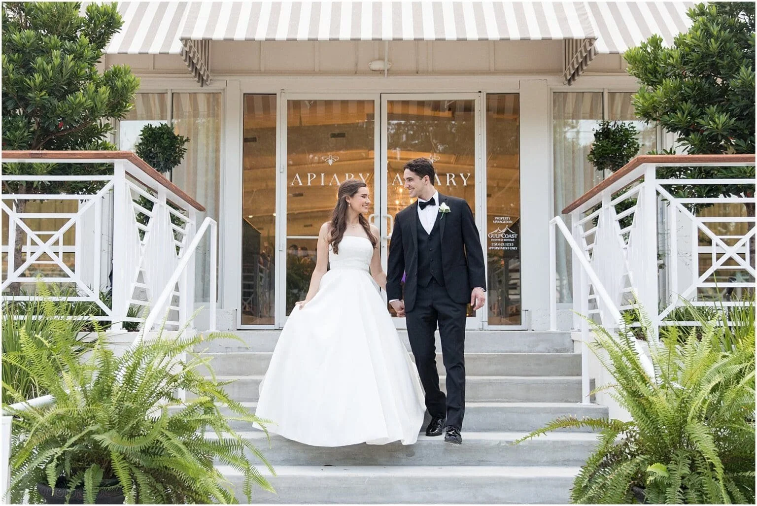 A bride and groom walking out of a wedding venue holding hands, surrounded by greenery and steps.