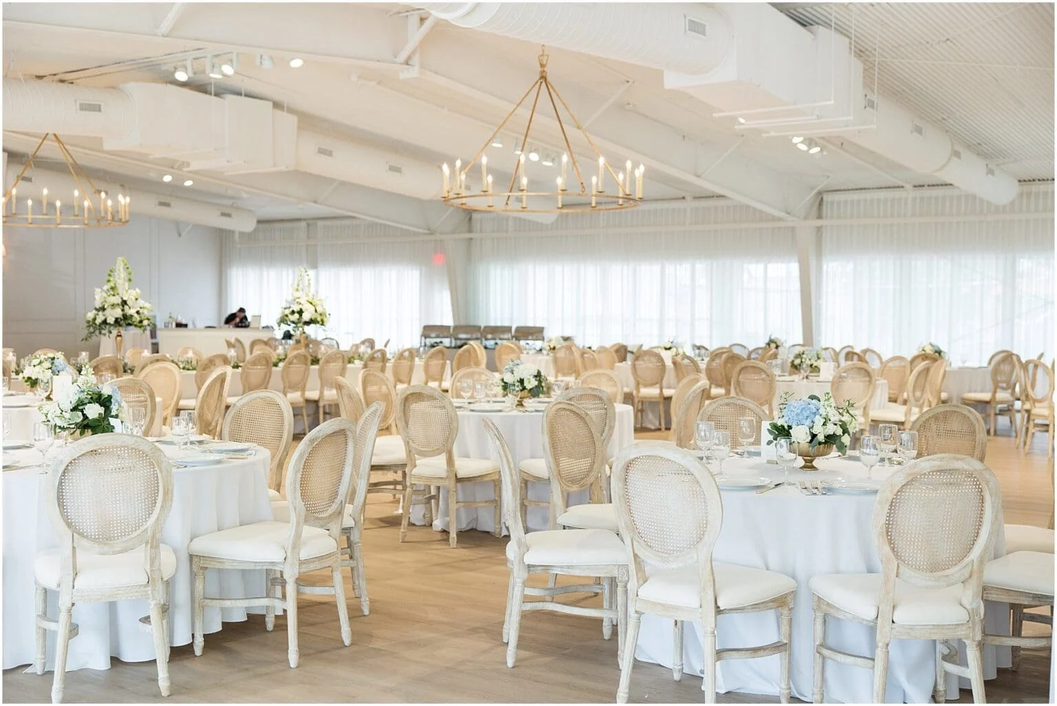Elegant event space decorated with round tables covered in white tablecloths, floral centerpieces with white and blue flowers, beige chairs with woven backs, brass chandeliers, and large windows with sheer curtains.