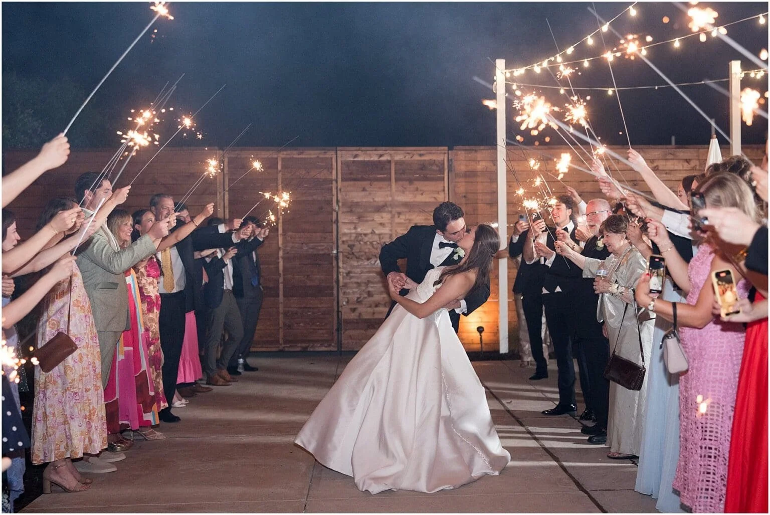 A bride and groom sharing a kiss while dancing at their wedding reception, surrounded by guests holding sparklers during nighttime.