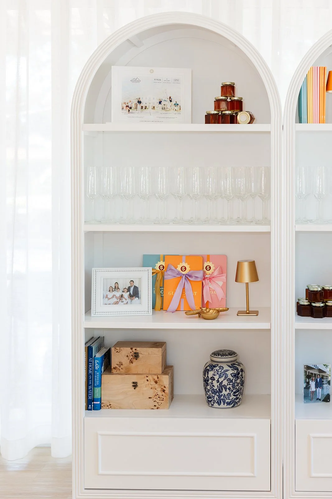 Decorative white bookshelf with framed family photo, gift boxes with ribbons, small gold lamp, jars, books, wooden boxes, and a blue and white floral vase.