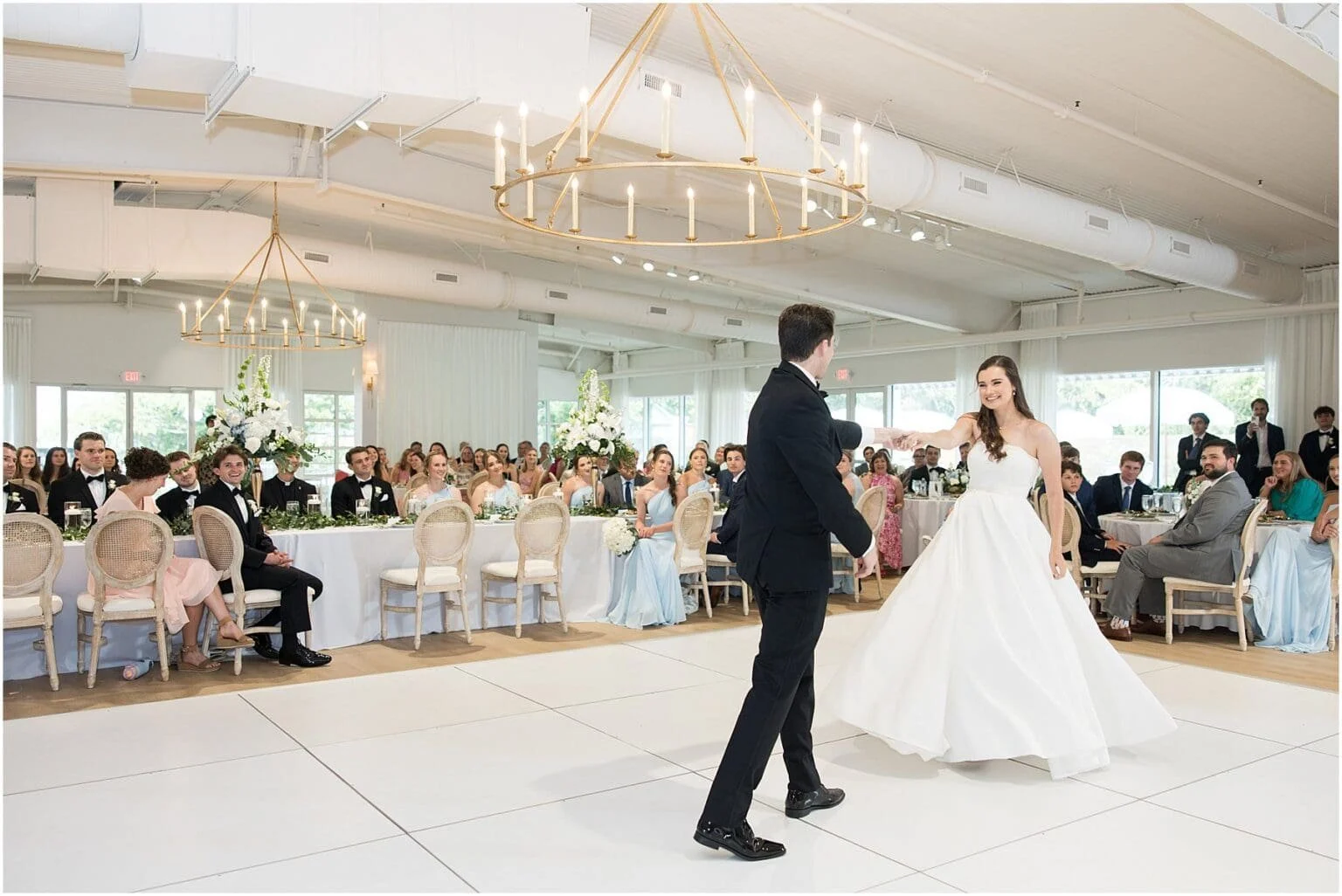 A bride and groom dancing at their wedding reception in a decorated banquet hall, with guests seated at tables watching and smiling.