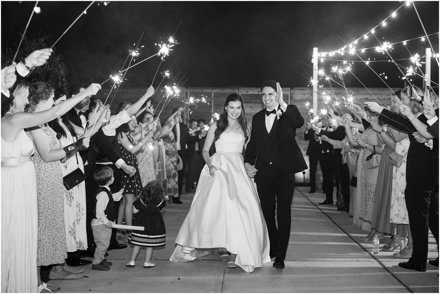 A black and white photo of a newlywed couple walking hand in hand through a crowd of wedding guests holding sparklers at night.