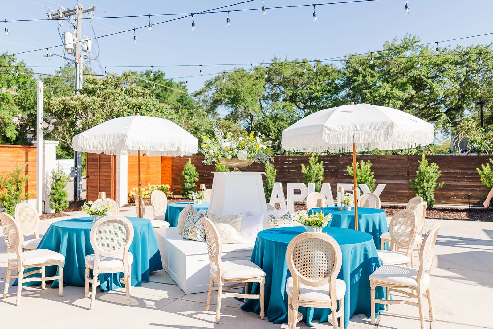 Outdoor event setup with round tables covered in blue tablecloths, white chairs, white umbrellas, and floral centerpieces with trees and string lights in the background.