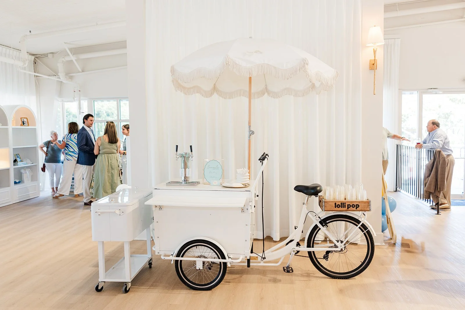 A white tricycle cart with a large white umbrella and a small sign that reads 'lolli pop', positioned indoors on a wooden floor with people gathered in the background.