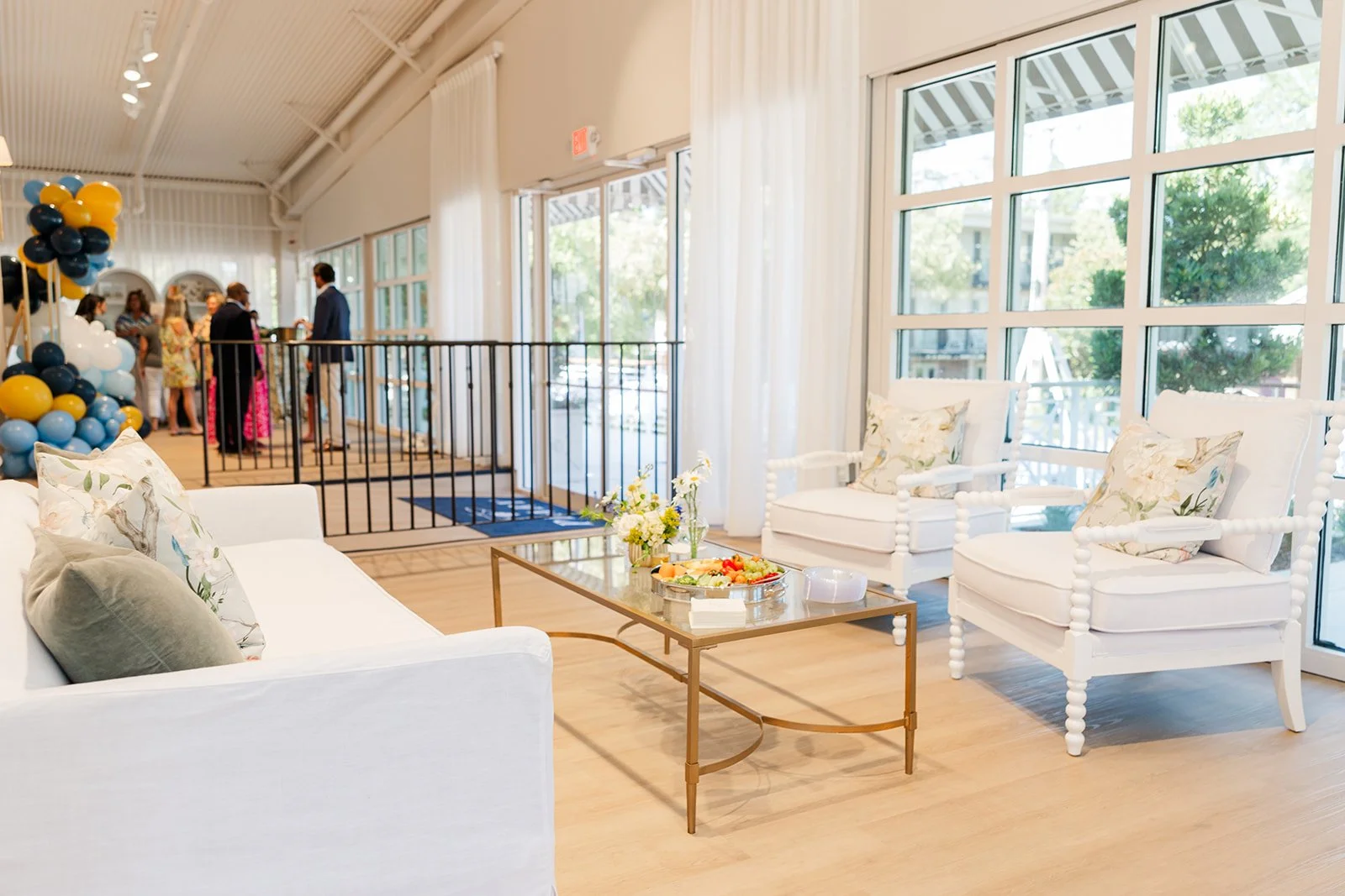 Brightly lit indoor space with white furniture, a glass coffee table with snacks and flowers, large windows, and a group of people socializing near balloons.