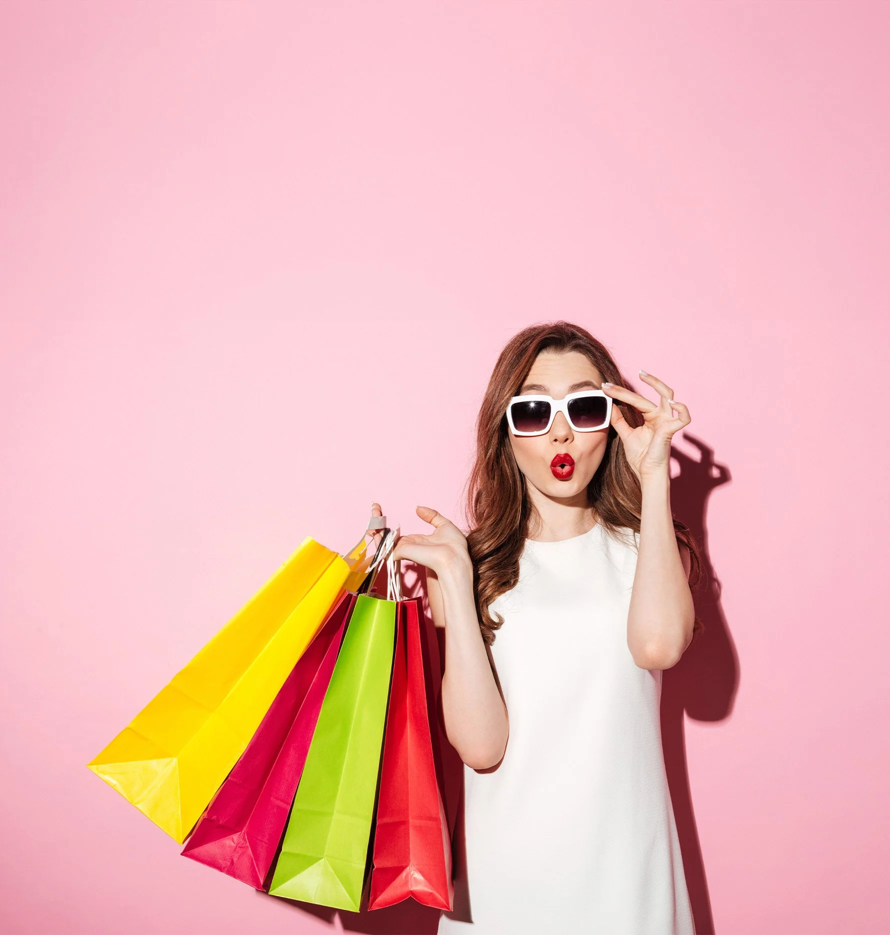 Woman holding colorful shopping bags and wearing sunglasses against a pink background.