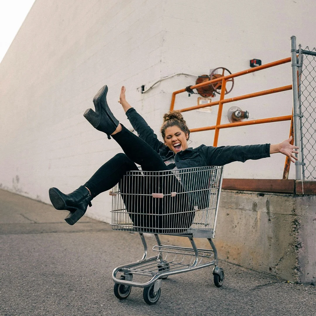 Person sitting joyfully in shopping cart on street with arms and legs outstretched, wearing denim outfit and black boots.