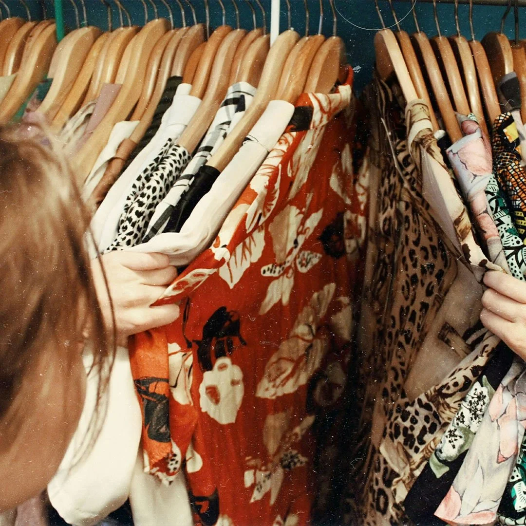 Person browsing clothes on wooden hangers in a closet, examining colorful garments with floral and leopard patterns.