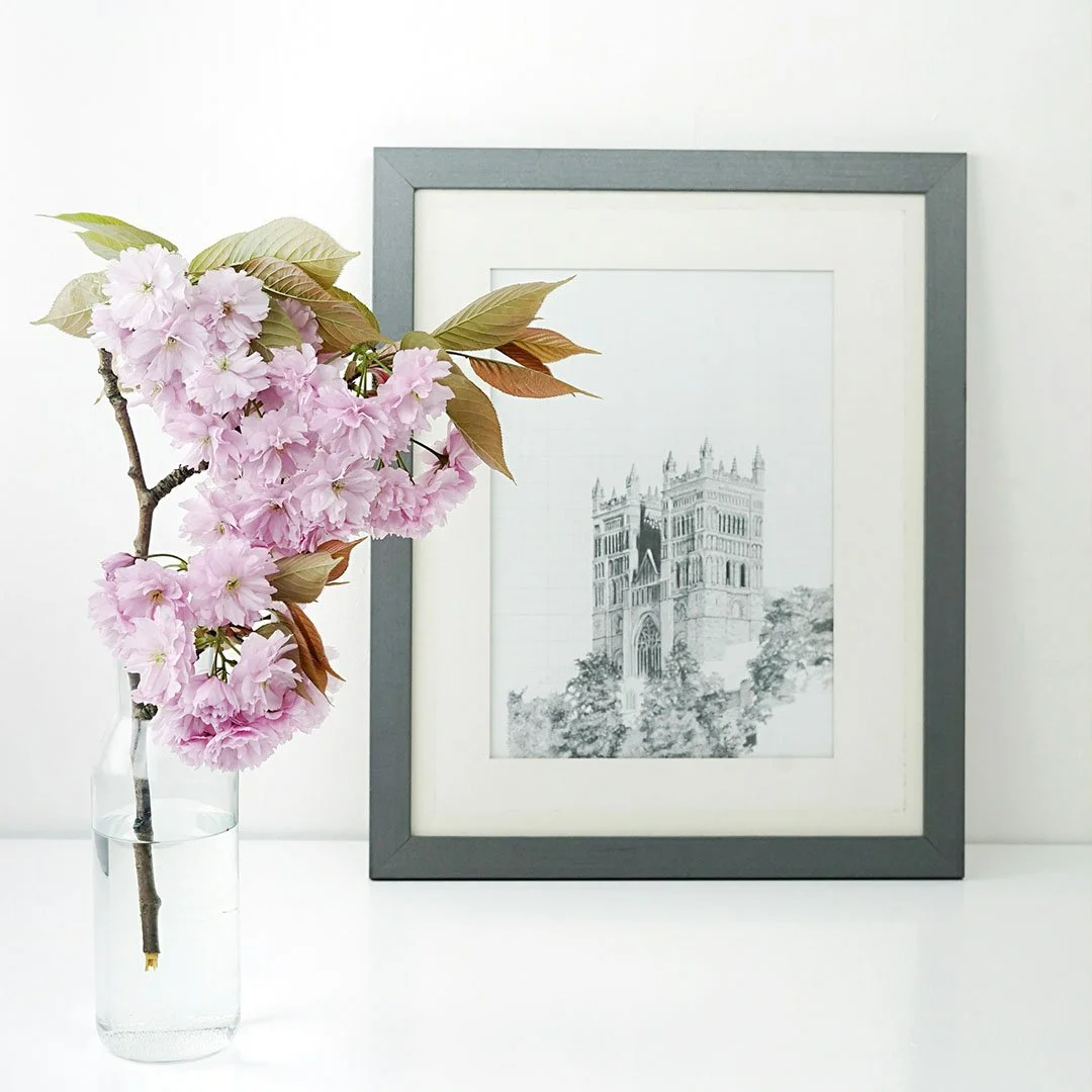 Cherry blossoms in a glass vase next to a framed architectural sketch of a historic building.