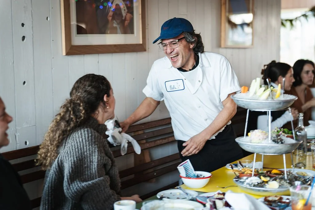 A man in a white shirt and blue cap laughing and talking to a woman with curly hair at a gathering, with a table of food and desserts in front of them.