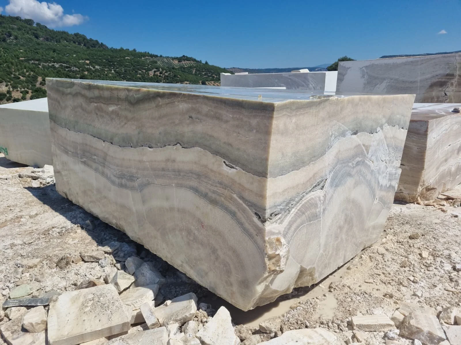 Large, rectangular Onyx block with natural green, beige, and gray veining, set outdoors on a dirt ground, with a scenic backdrop of hills and clear blue sky.