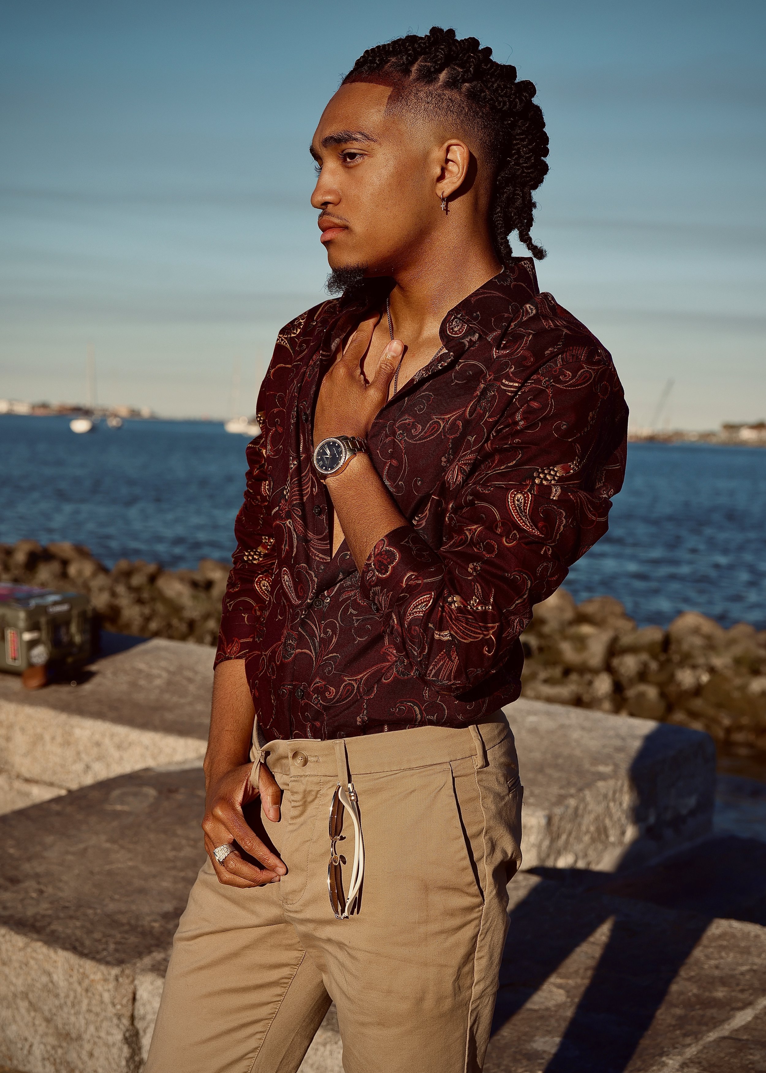 Man with braided hairstyle wearing a patterned shirt and beige pants, standing by a waterfront.
