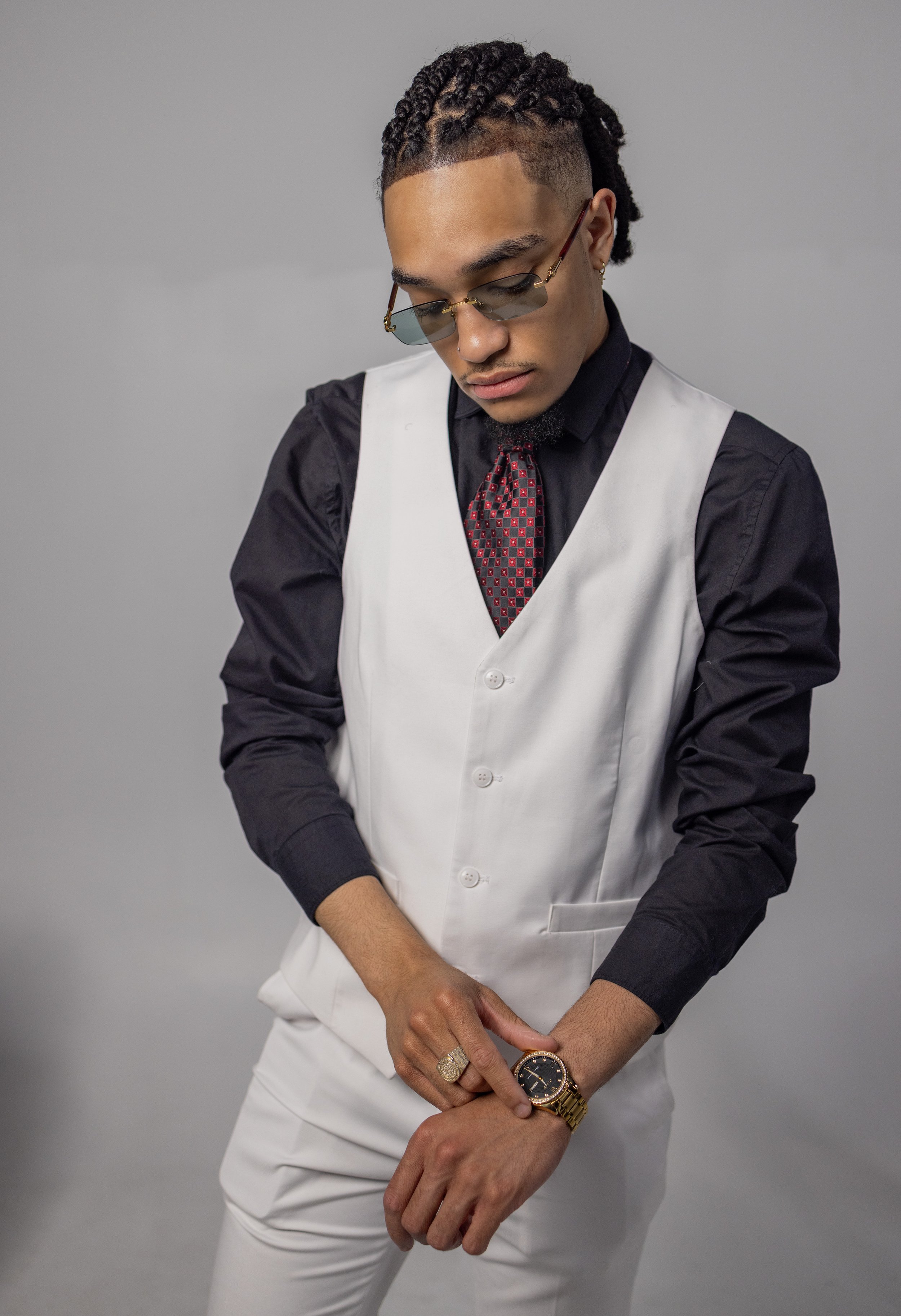 A man wearing black sunglasses, a black shirt, a white vest, and white pants, with braided hair, checking his gold wristwatch.