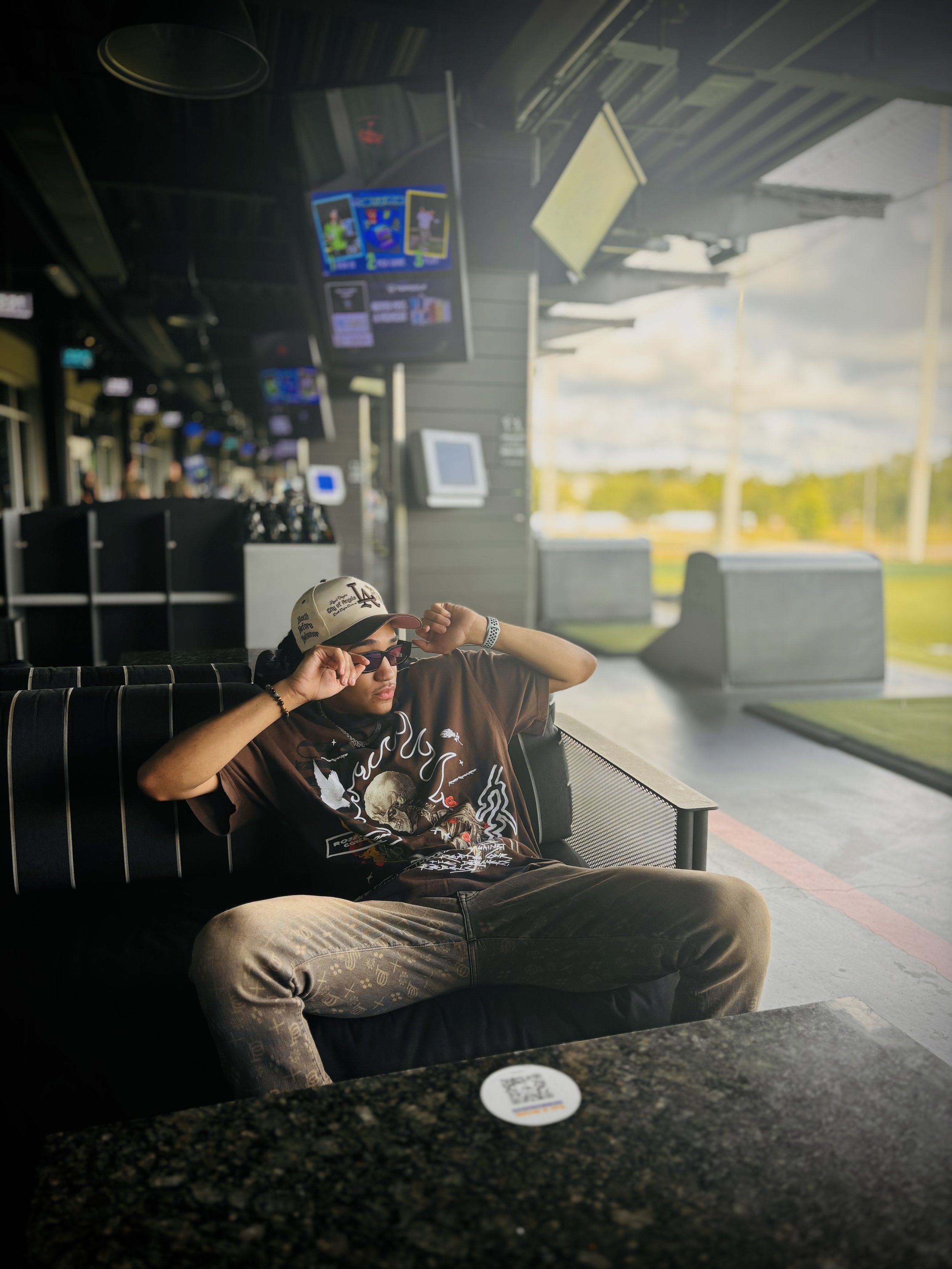 Person sitting on a couch at a golf entertainment venue with TV screens and golf bays in the background.