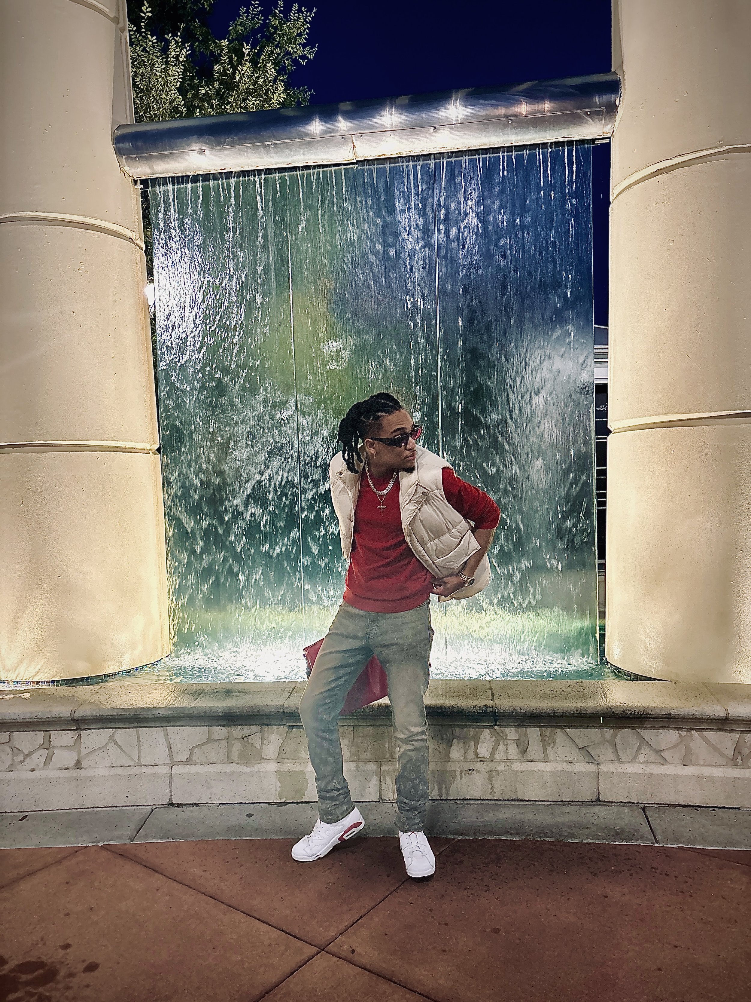 Person posing in front of a decorative waterfall, wearing a red shirt, white puffy vest, jeans, and white sneakers.