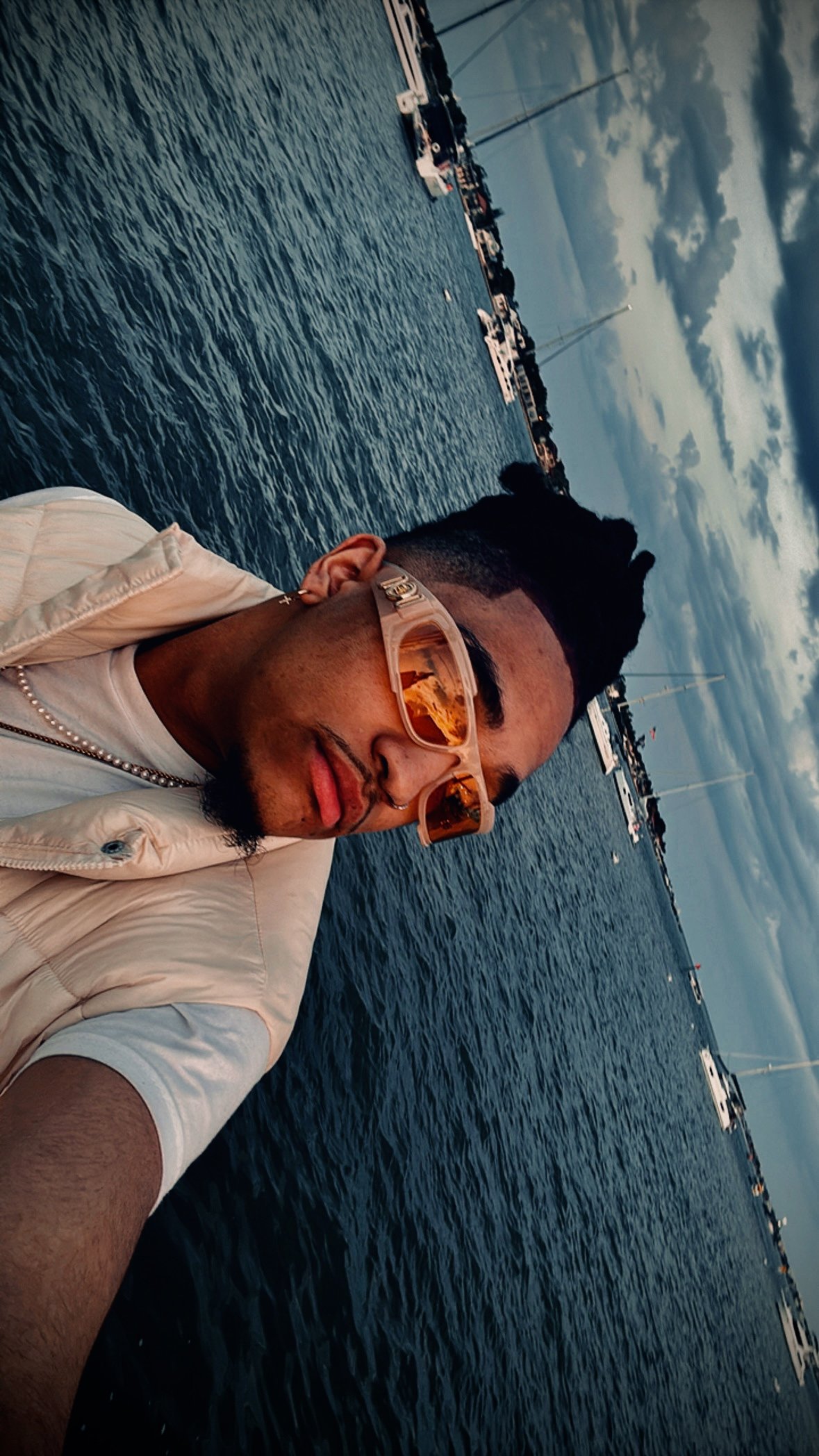 Person in sunglasses posing near water with boats in the background.