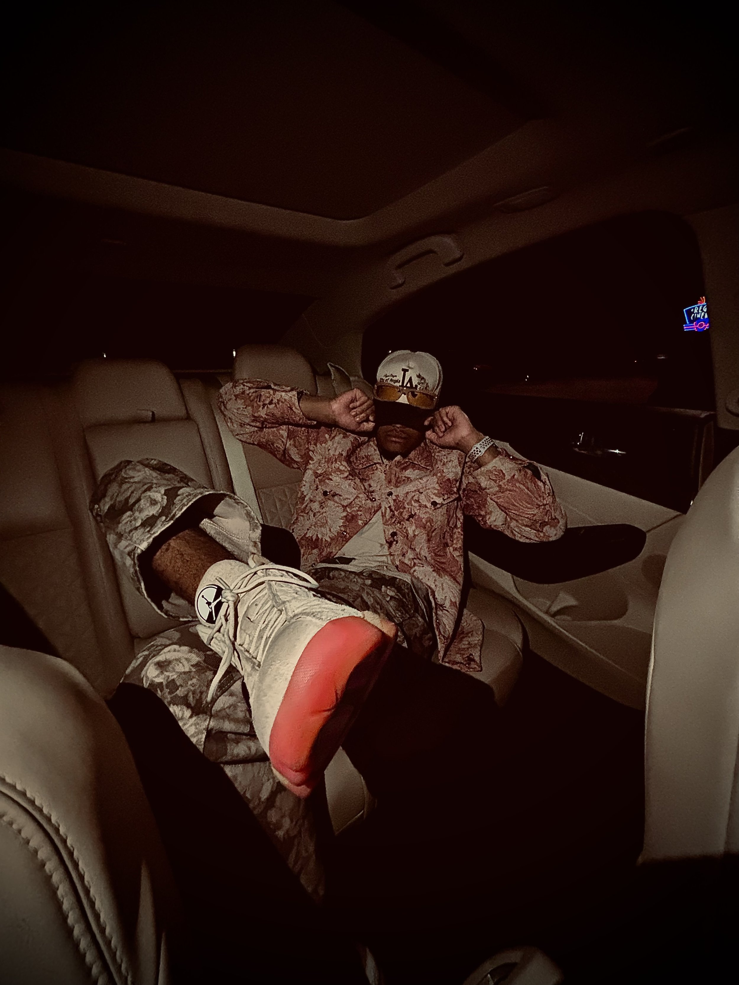 Person reclining in a car's backseat wearing a patterned shirt, camouflage pants, white sneakers with red soles, and a cap with 'LA' on it.