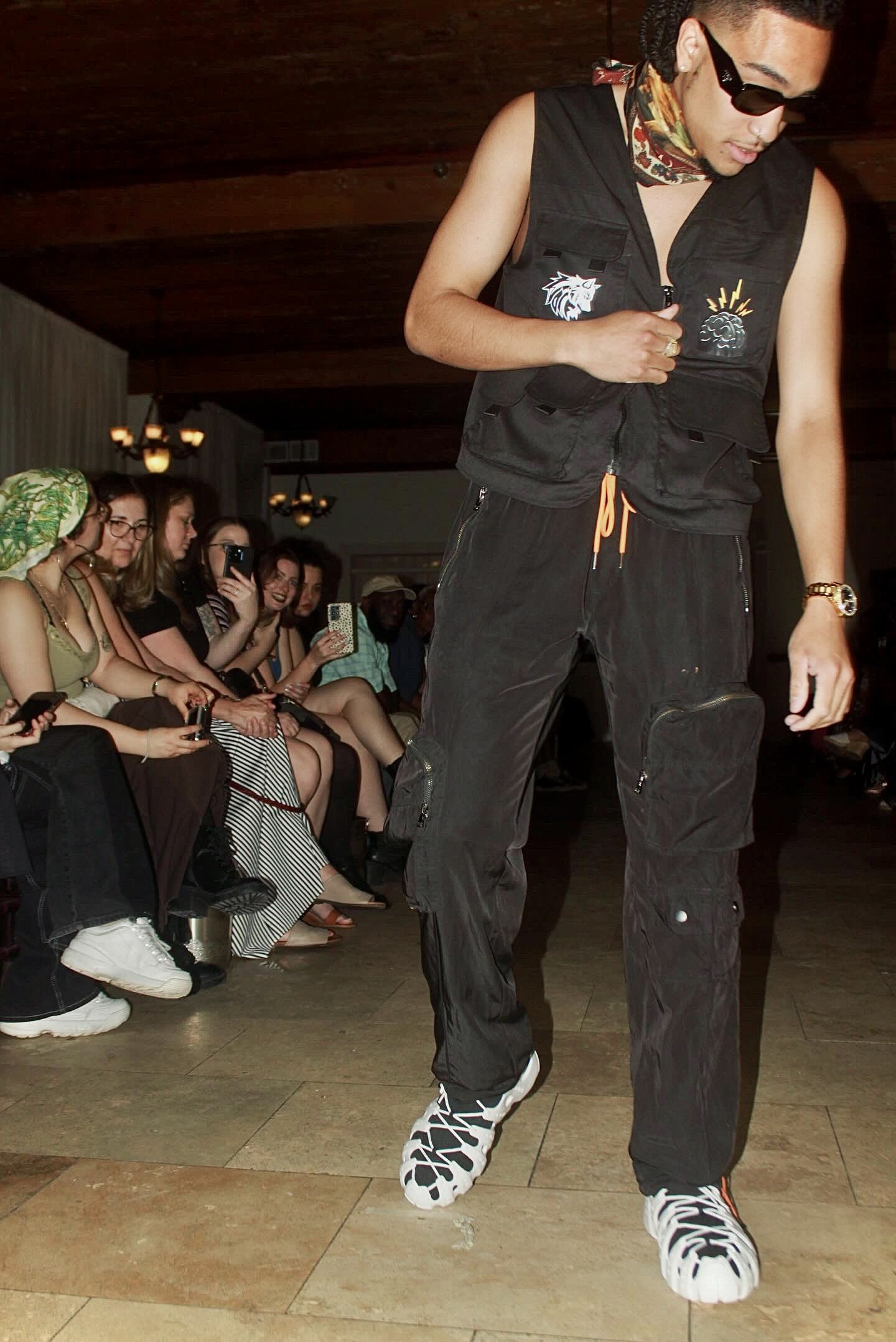 Fashion show with a model wearing a black vest and pants, white sneakers, and sunglasses, walking in front of seated audience snapping photos.