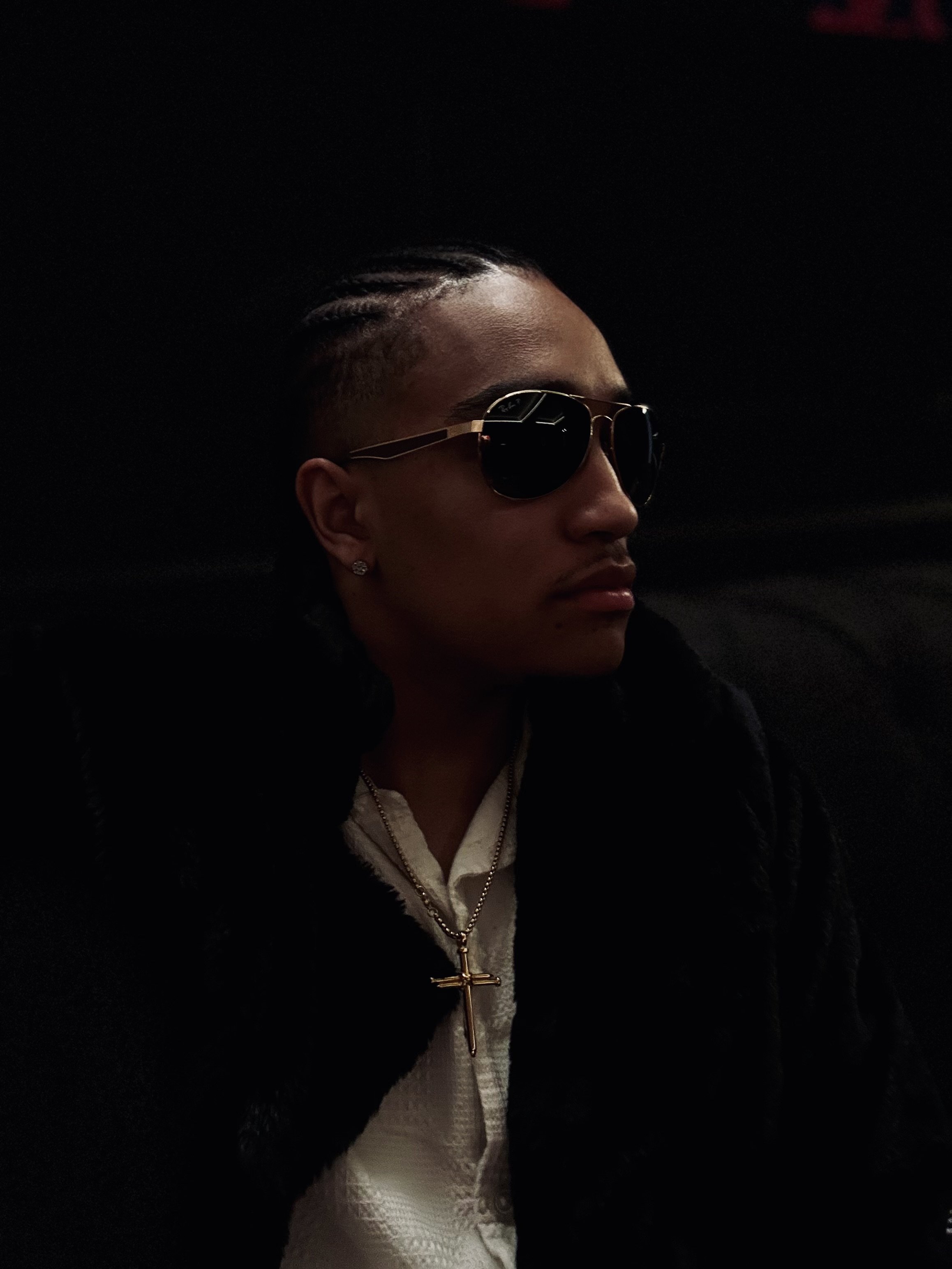 Person wearing sunglasses and a fur coat, with a cross necklace, sitting in a dimly lit environment.