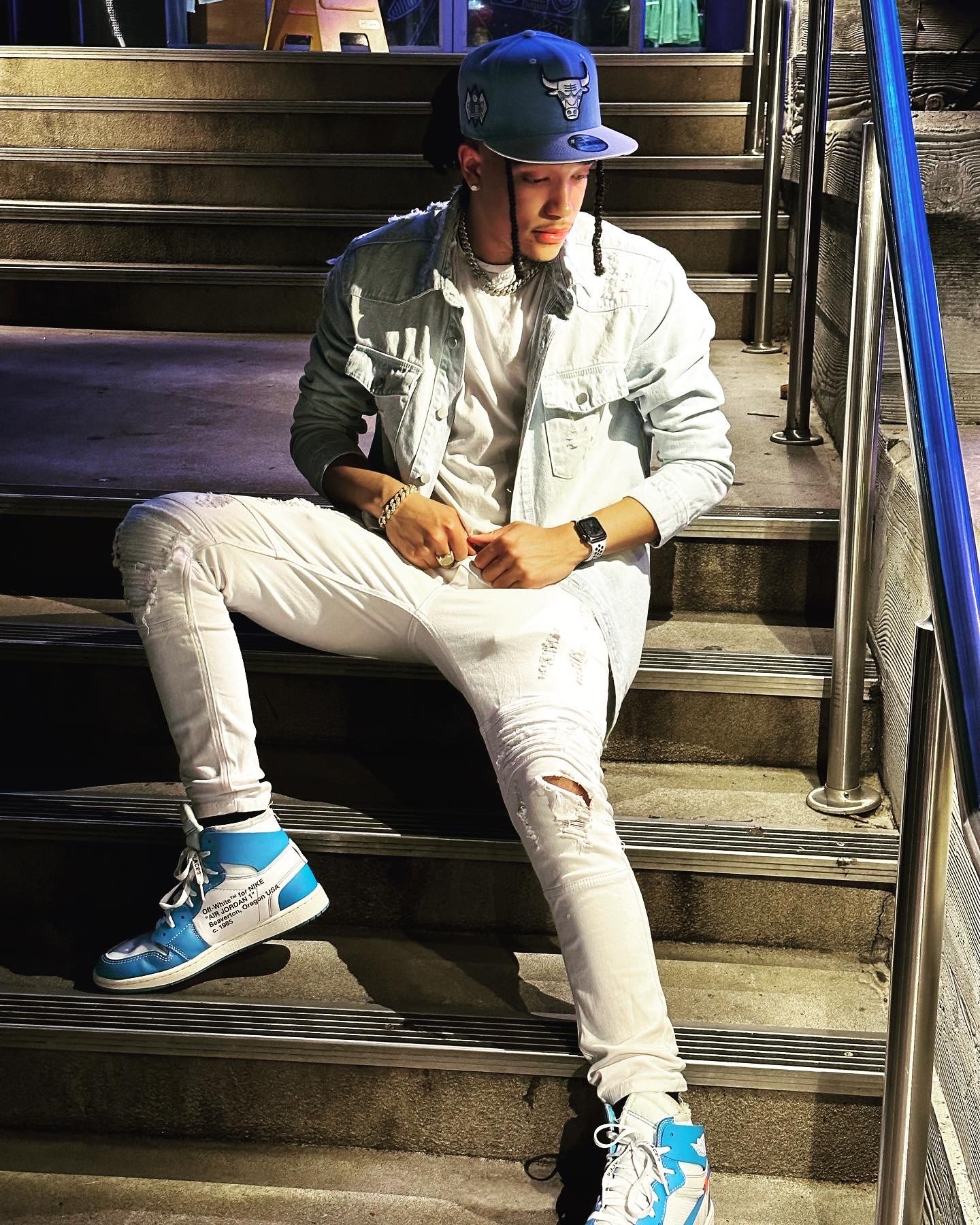 A person sitting on steps wearing a blue baseball cap, light denim jacket, white shirt, ripped white jeans, and blue and white sneakers.
