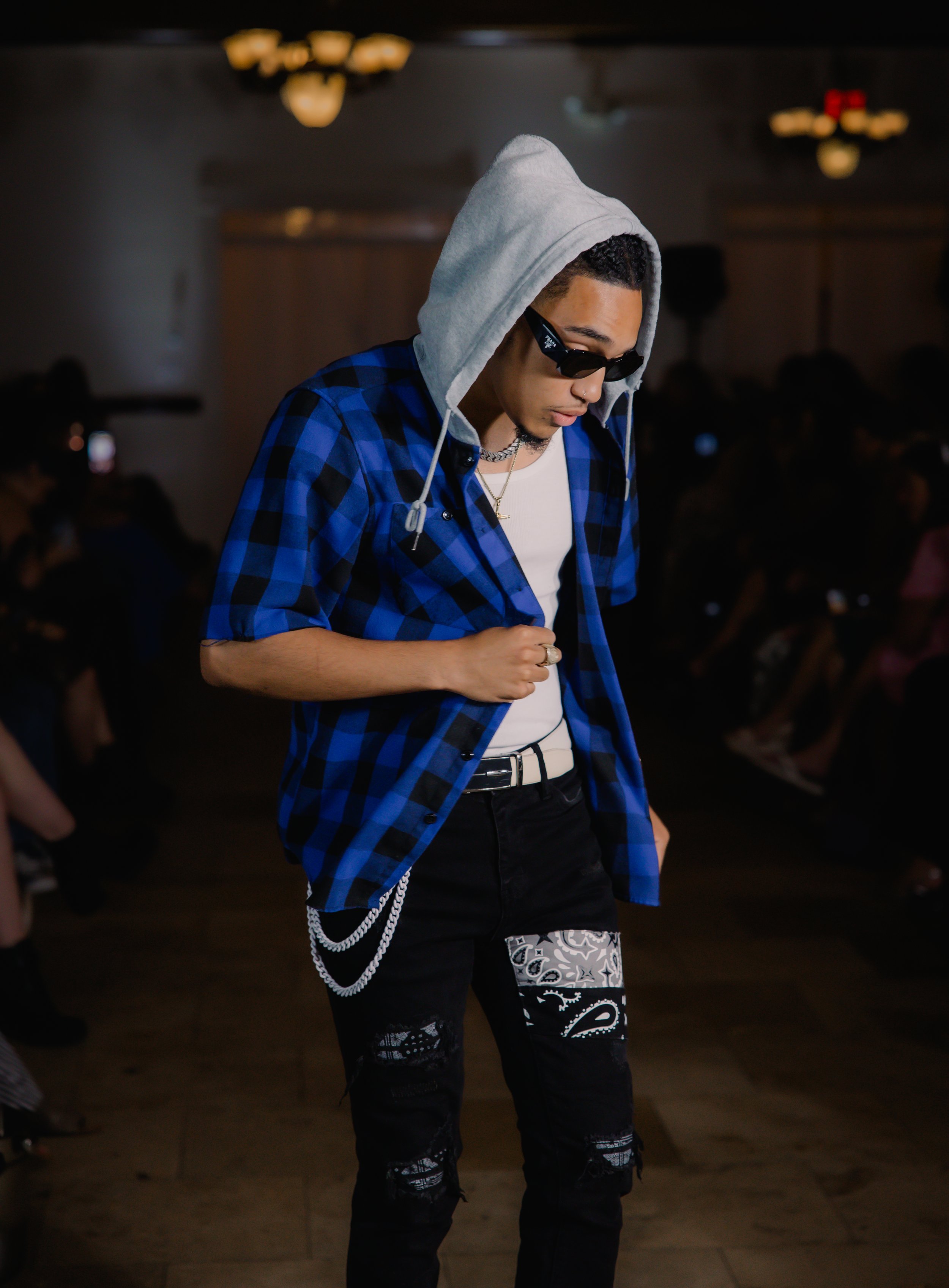 Person wearing a blue and black checkered shirt, gray hood, black sunglasses, white tank top, and black jeans with patterns, walking in a dimly lit room.