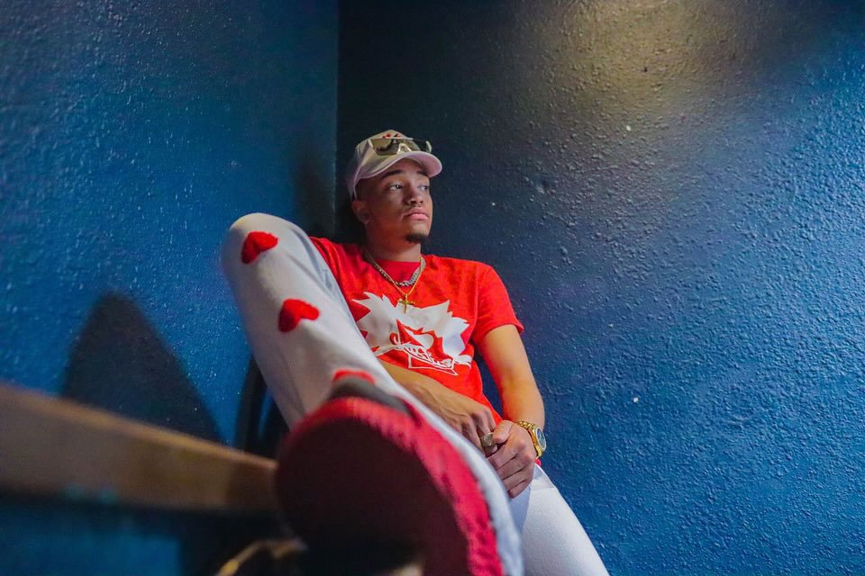 Person in a red shirt and white pants sitting against a dark blue wall, wearing a cap and looking relaxed.
