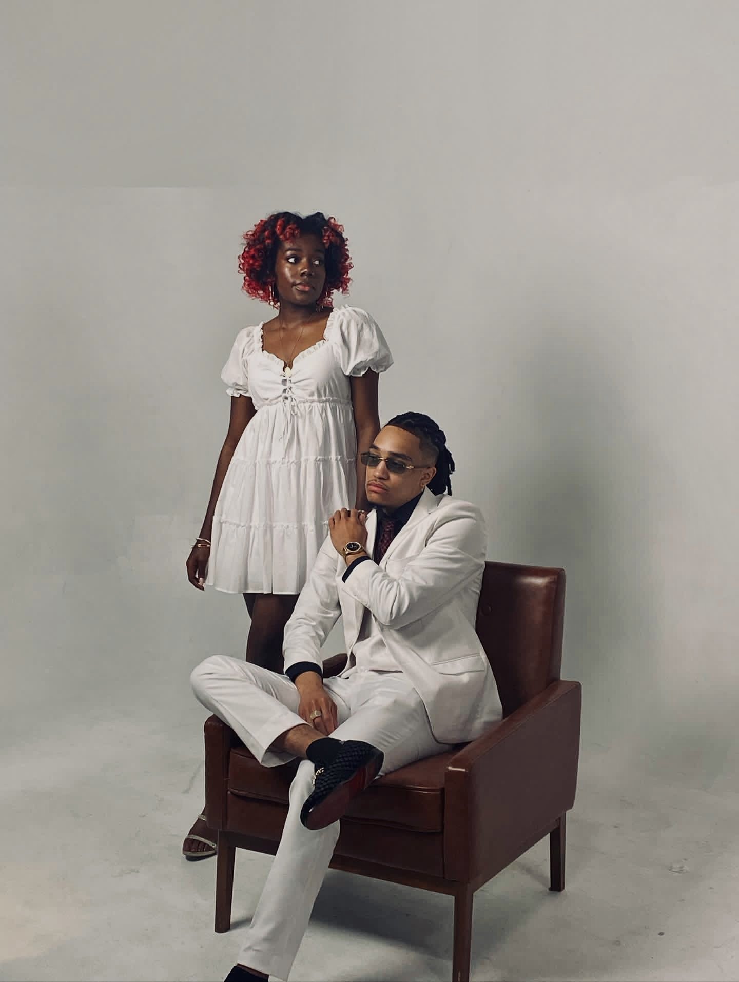 A woman in a white dress stands beside a seated man in a white suit and sunglasses. The man is sitting on a brown leather chair in a studio setting.