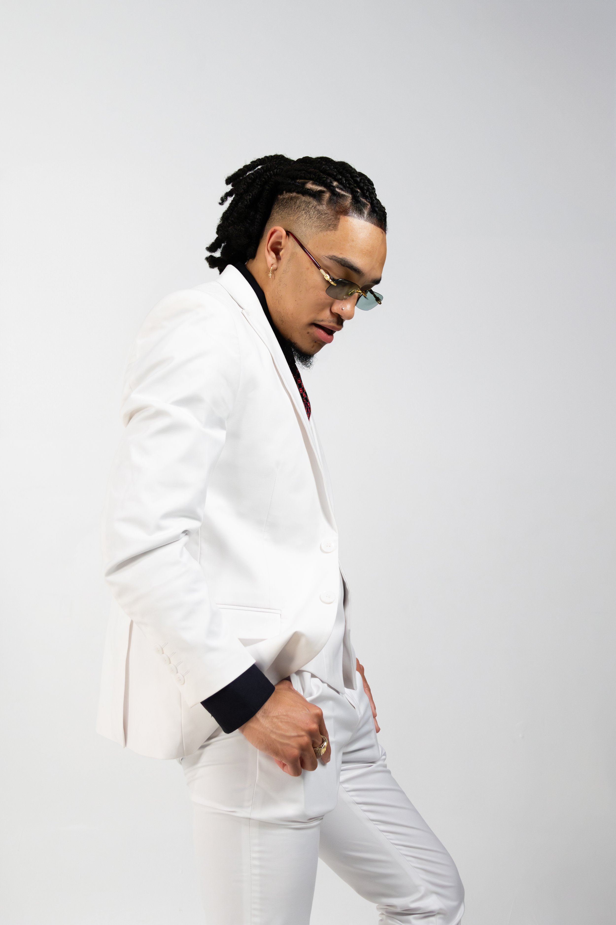 Man in white suit with braids and glasses, posing against a plain background.