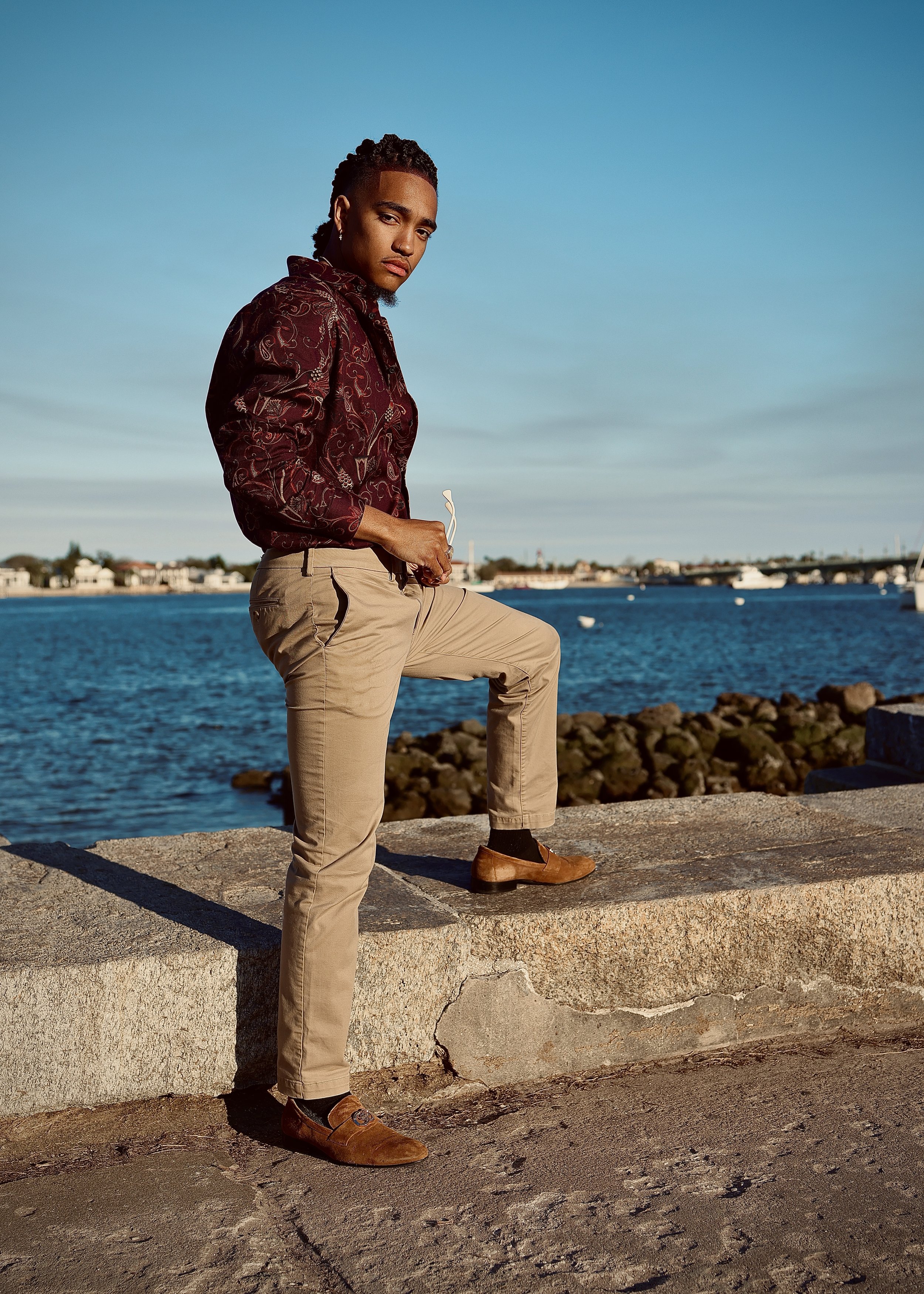 Person in stylish clothing standing by the waterfront with city skyline and bridge in the background.