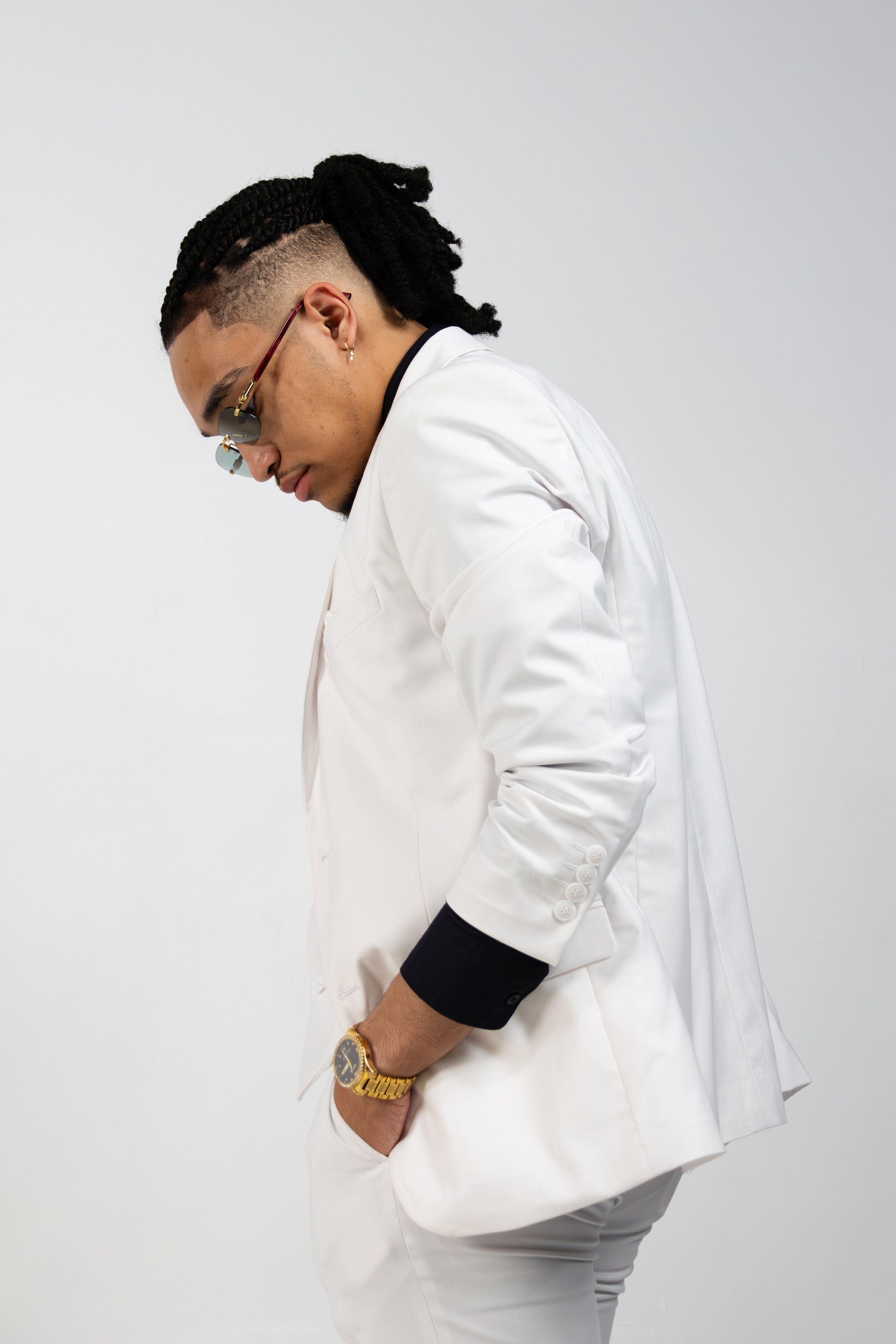 Person in a white suit with dreadlocks and sunglasses, looking downwards.