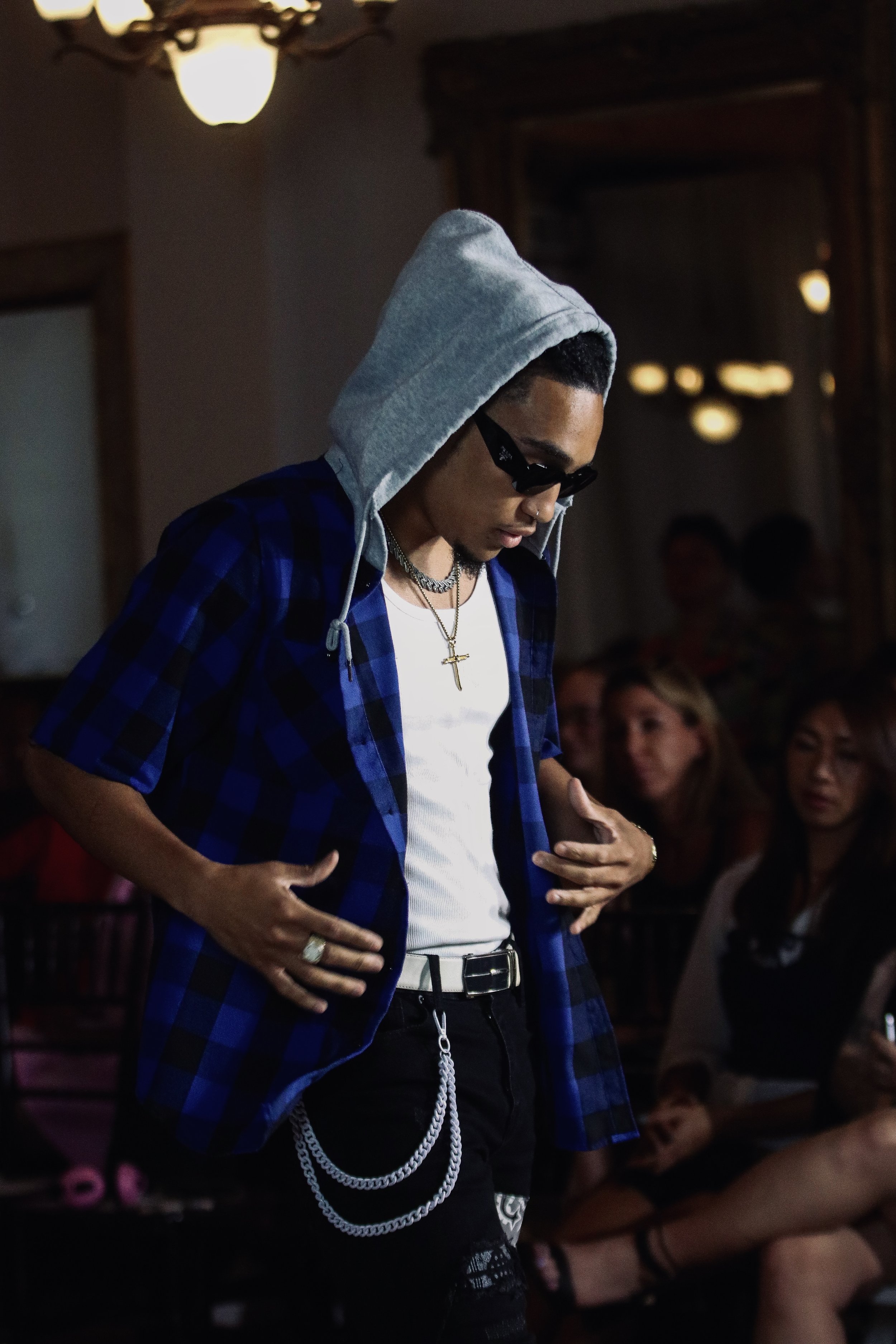 Person wearing a blue checkered shirt with a gray hood, black sunglasses, necklaces, and accessories, walking in a dimly lit room.