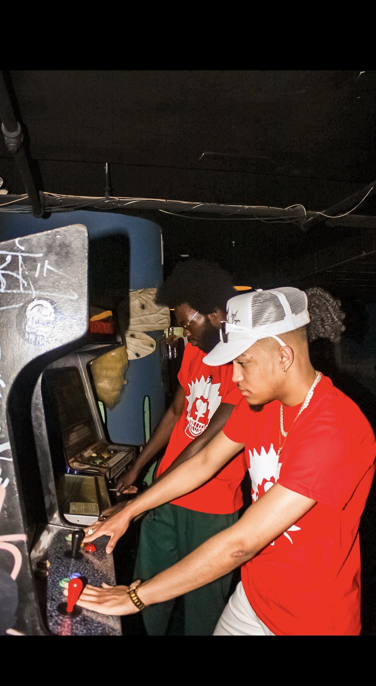 Two people in red shirts playing vintage arcade games in a dimly lit room.