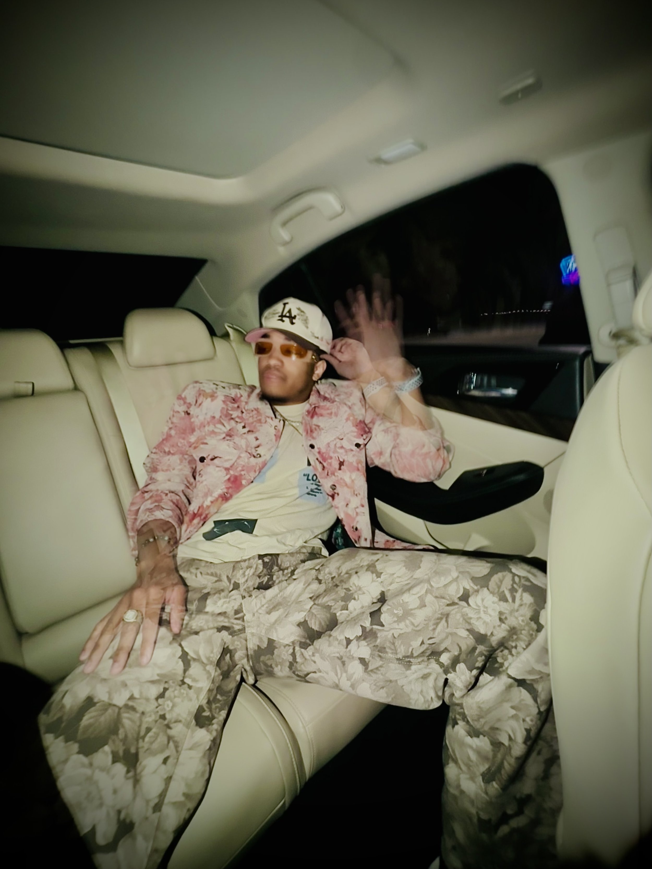 Person in floral outfit and baseball cap sitting in backseat of car