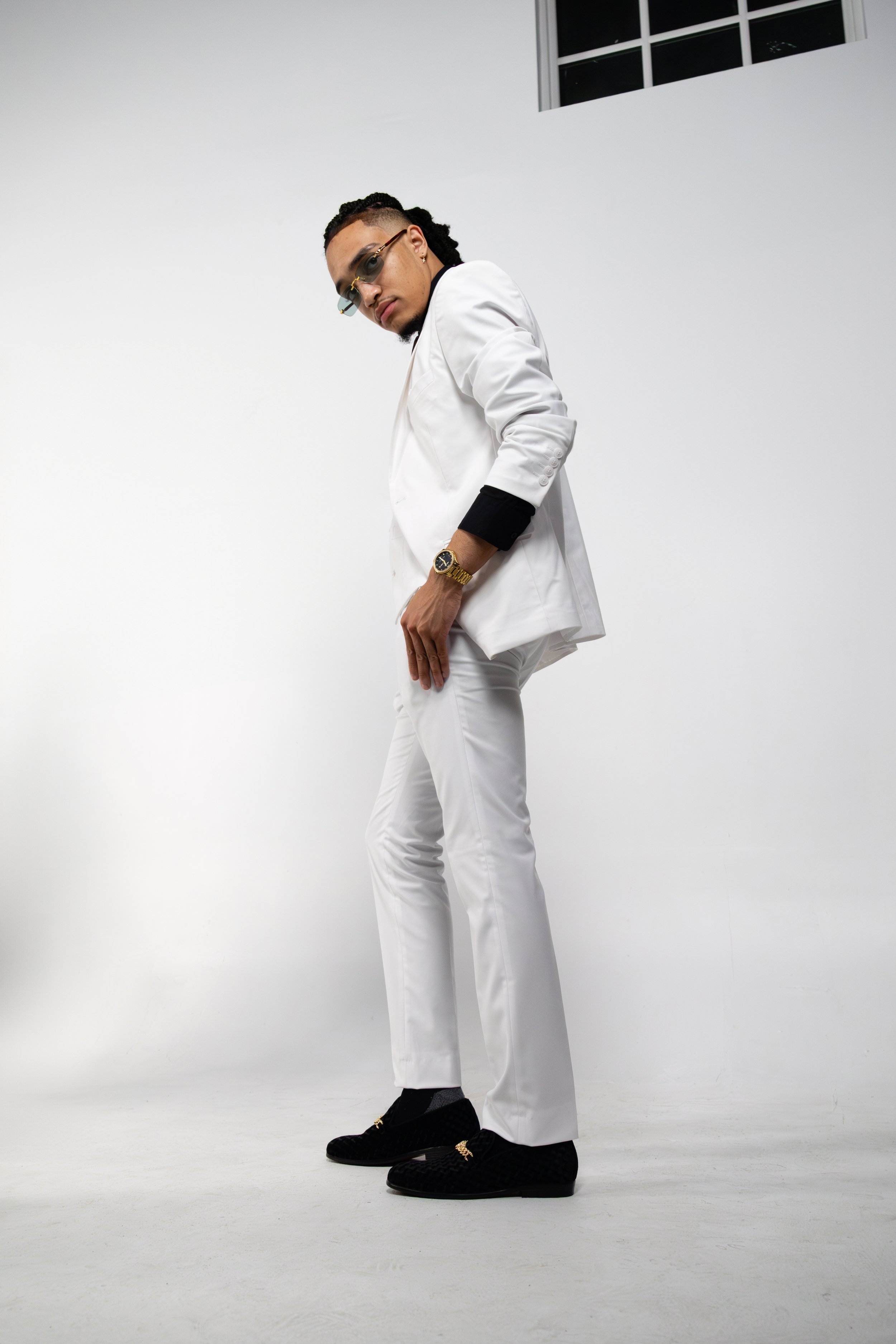 Person in a stylish white suit with black loafers and gold watch, posing in a minimalist white studio.