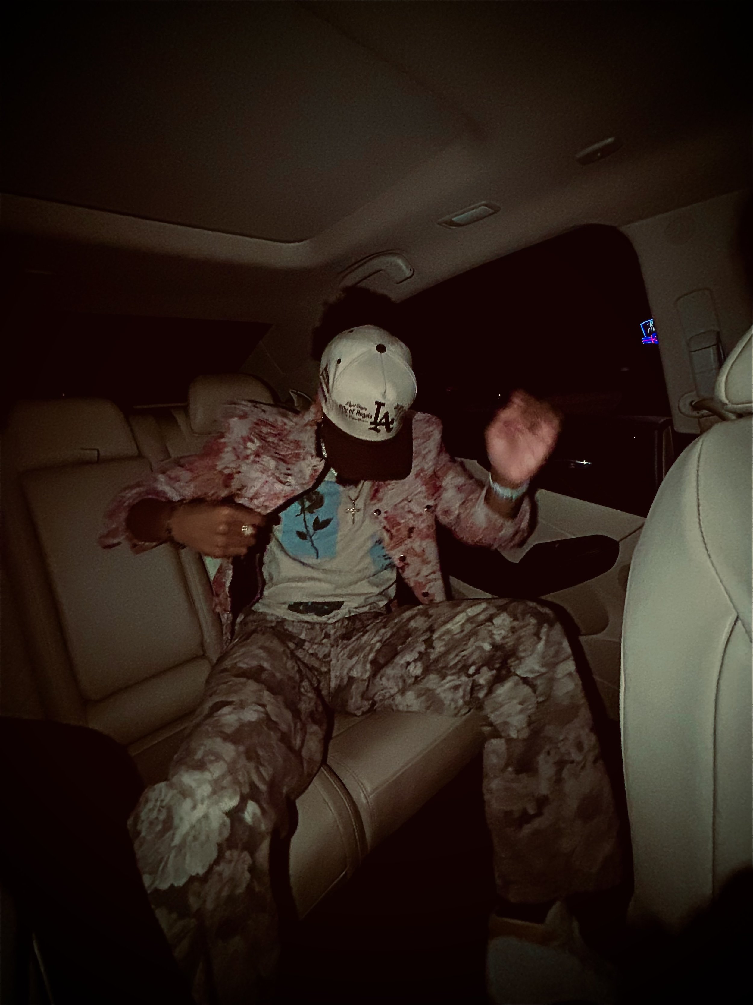 Person sitting in a car wearing a baseball cap, pink jacket, patterned pants, and a T-shirt.