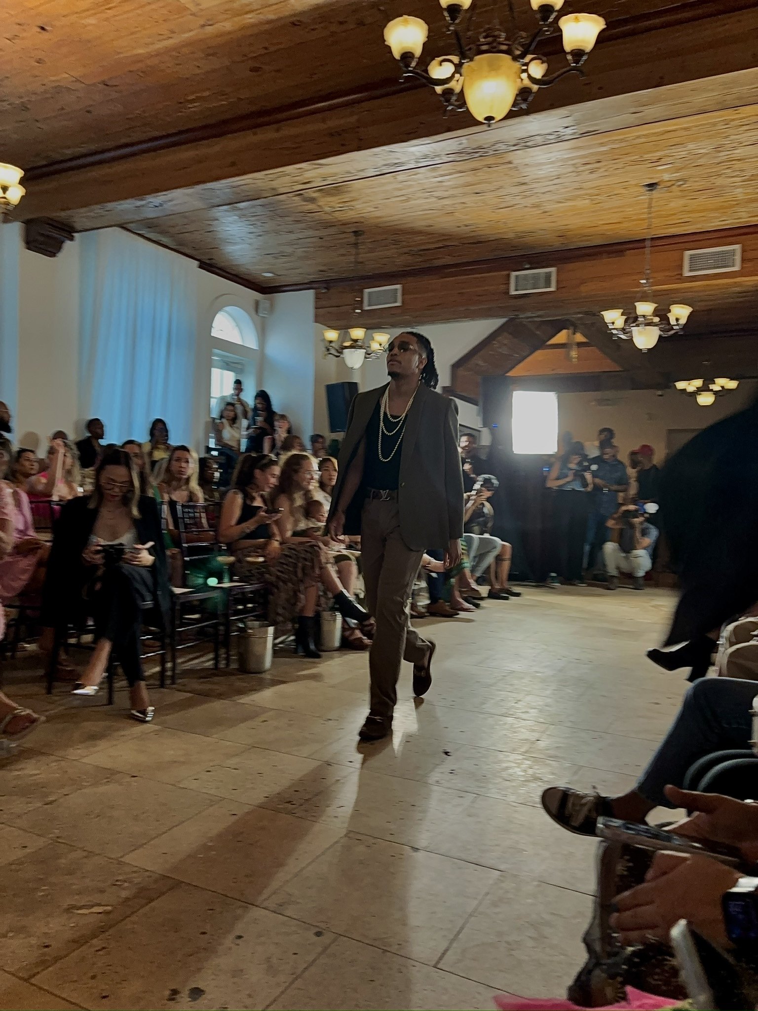 Model walking down a runway in a formal fashion show, wearing a suit and accessorized with necklaces, surrounded by seated audience members in a well-lit room with wooden ceilings and chandeliers.